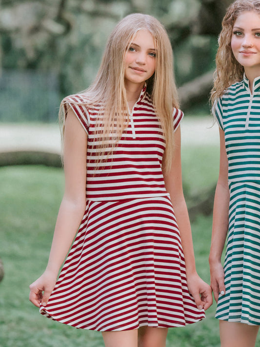 Model outside in wooded green area wearing a crimson and ivory striped dress with white canvas shoes. Model is slightly holding out the side of the dress. There is another girl beside her on the right wearing a green and ivory striped dress too.