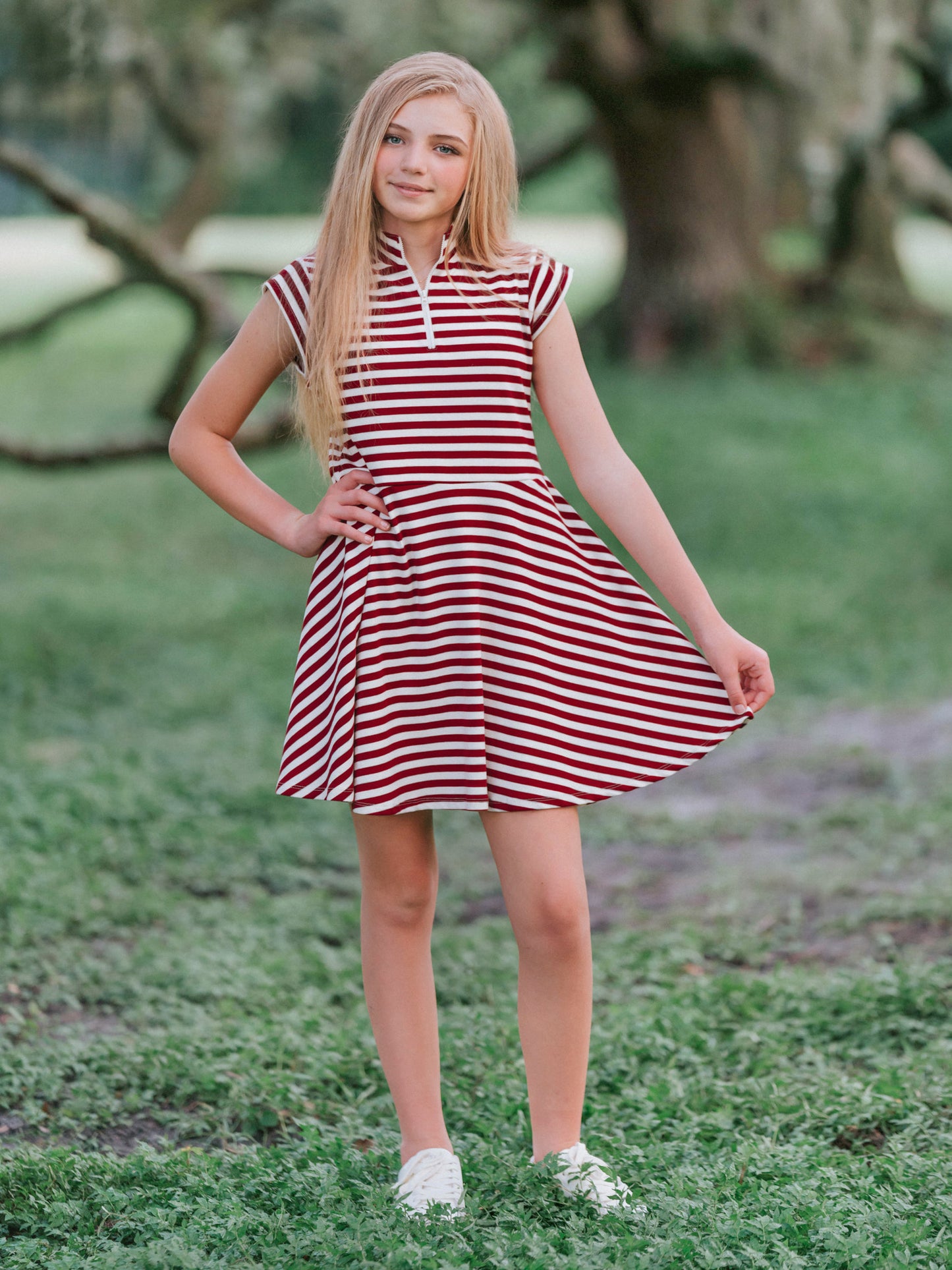 Model outside in wooded green area wearing a crimson and ivory striped dress with white canvas shoes. Model is slightly holding out the side of the dress. 