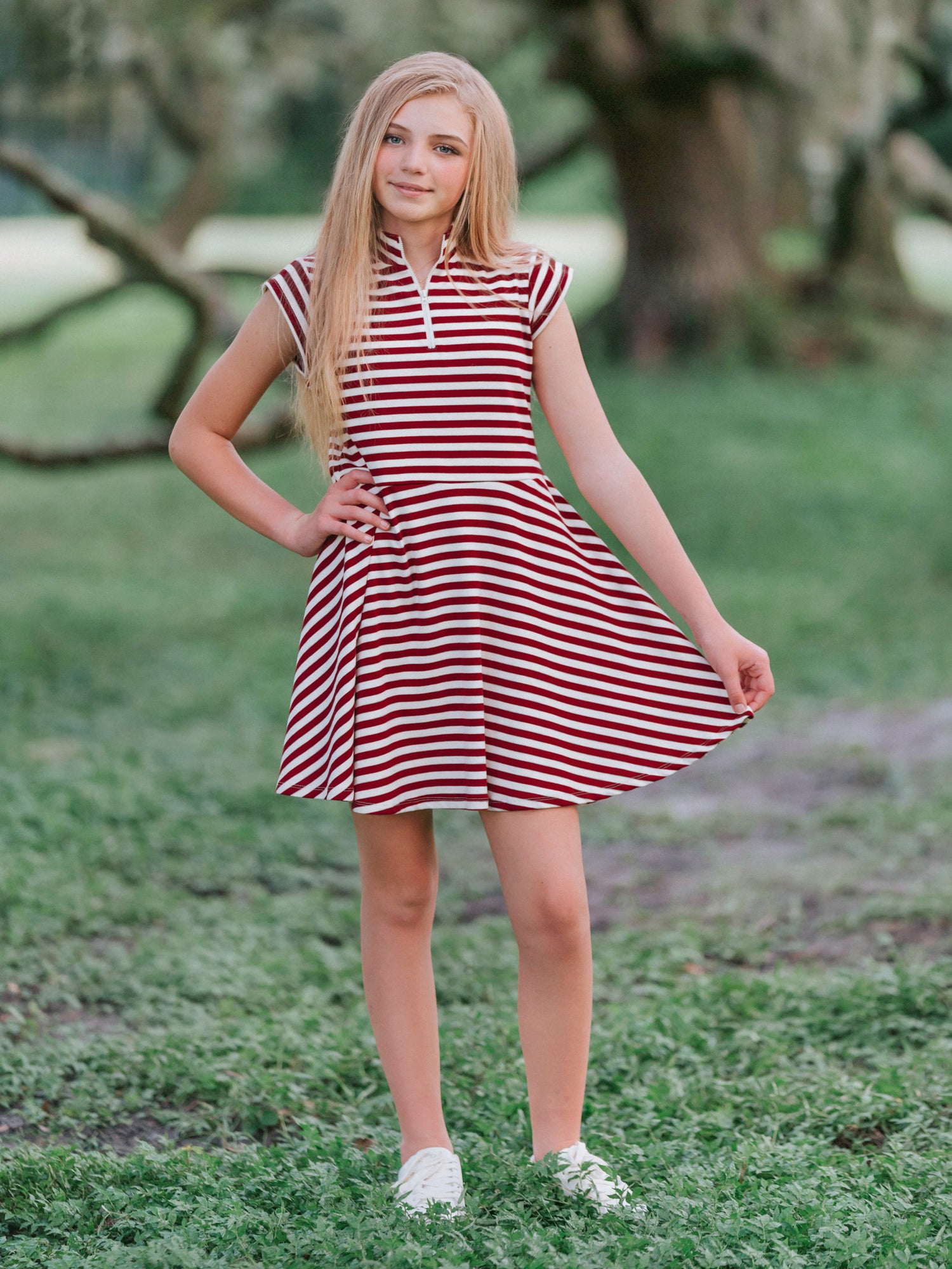Model outside in wooded green area wearing a crimson and ivory striped dress with white canvas shoes. Model is slightly holding out the side of the dress. 