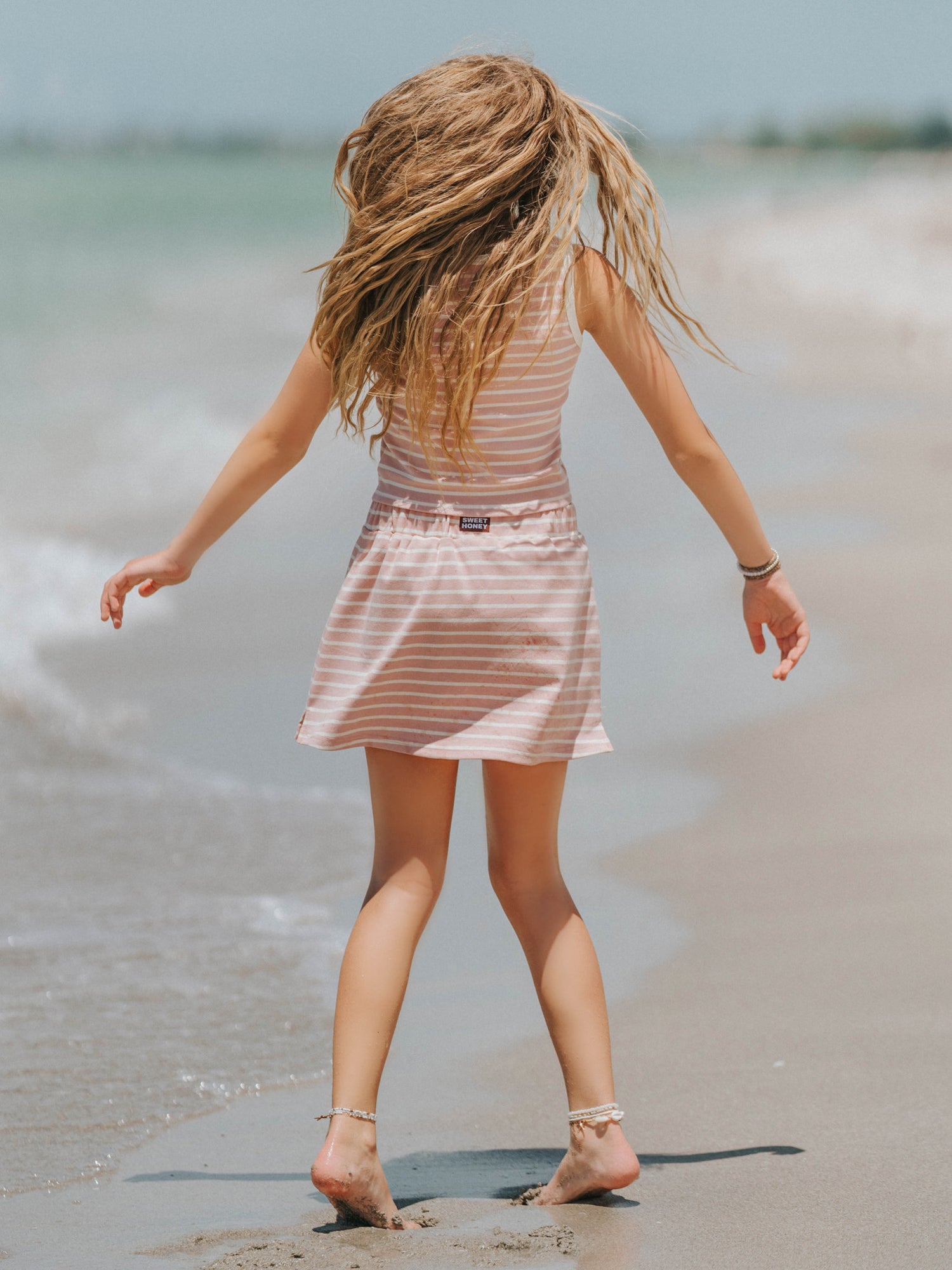 Model is outside on a beach wearing a two-piece matching pink and ivory striped skort set. Top is sleeveless. There is a SweetHoney logo tag on the back of the skort waistband