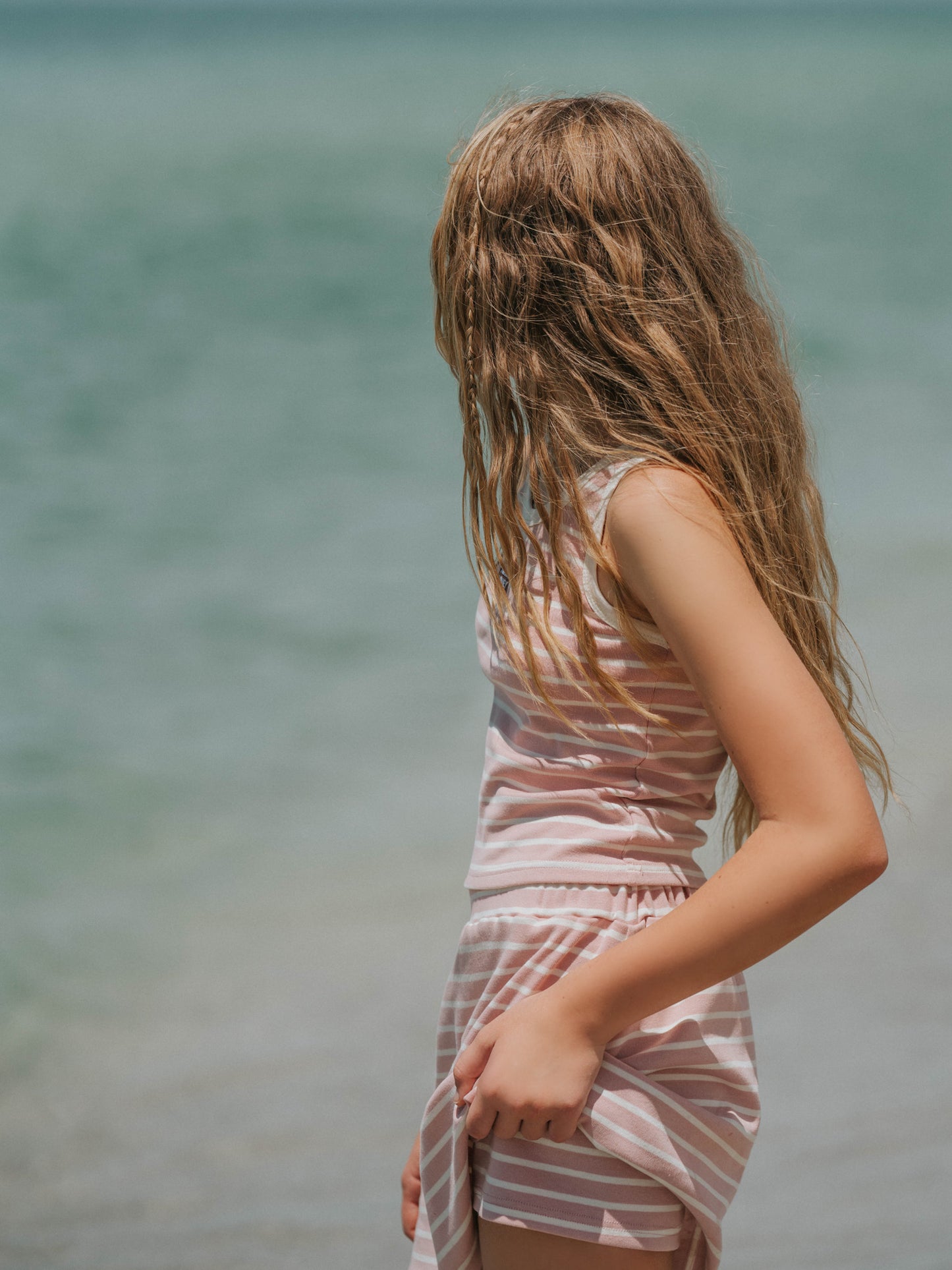Model is outside on a beach wearing a two-piece matching pink and ivory striped skort set. Top is sleeveless. Model is slightly holding up her skort to show the shorts underneath.
