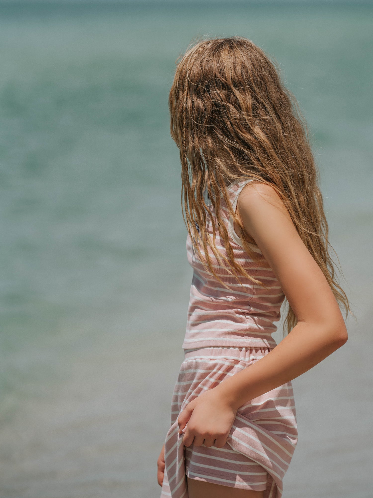 Model is outside on a beach wearing a two-piece matching pink and ivory striped skort set. Top is sleeveless. Model is slightly holding up her skort to show the shorts underneath.