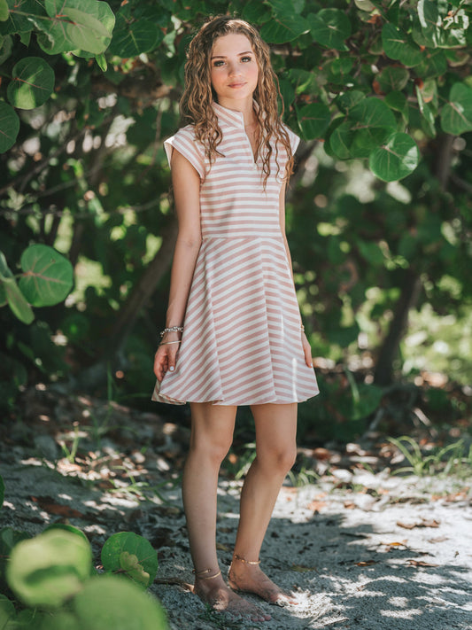 Model outside in a greenery area looking at the camera and wearing a dove pink and ivory striped dress. 