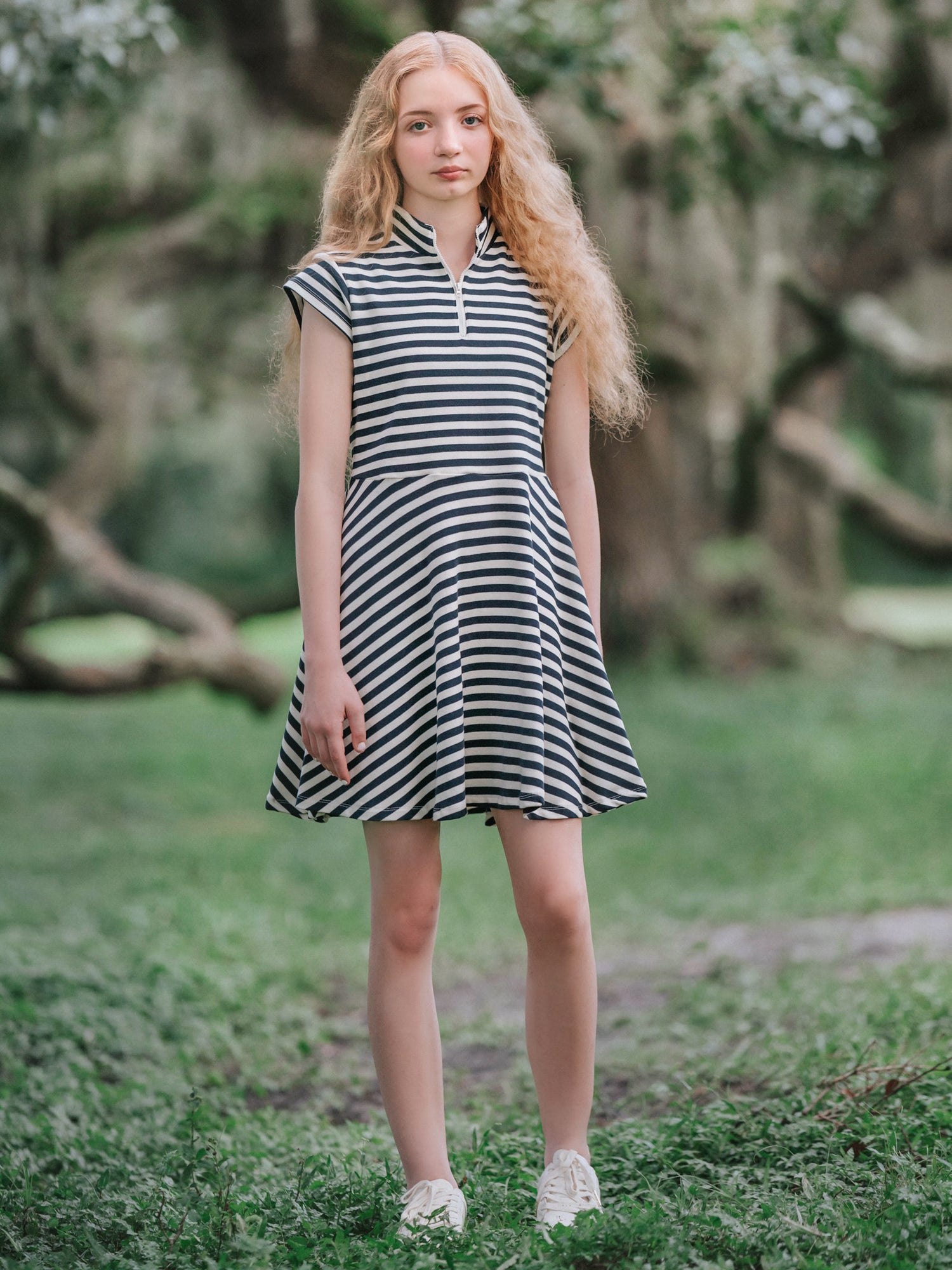 Girl outside in a mossy green area wearing hunter green and ivory striped dress and in white shoes. 