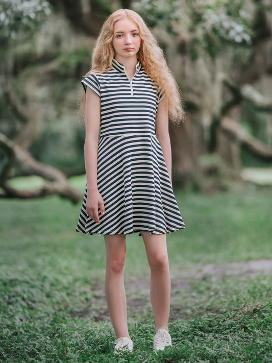 Girl outside in a mossy green area wearing hunter green and ivory striped dress and in white shoes. 