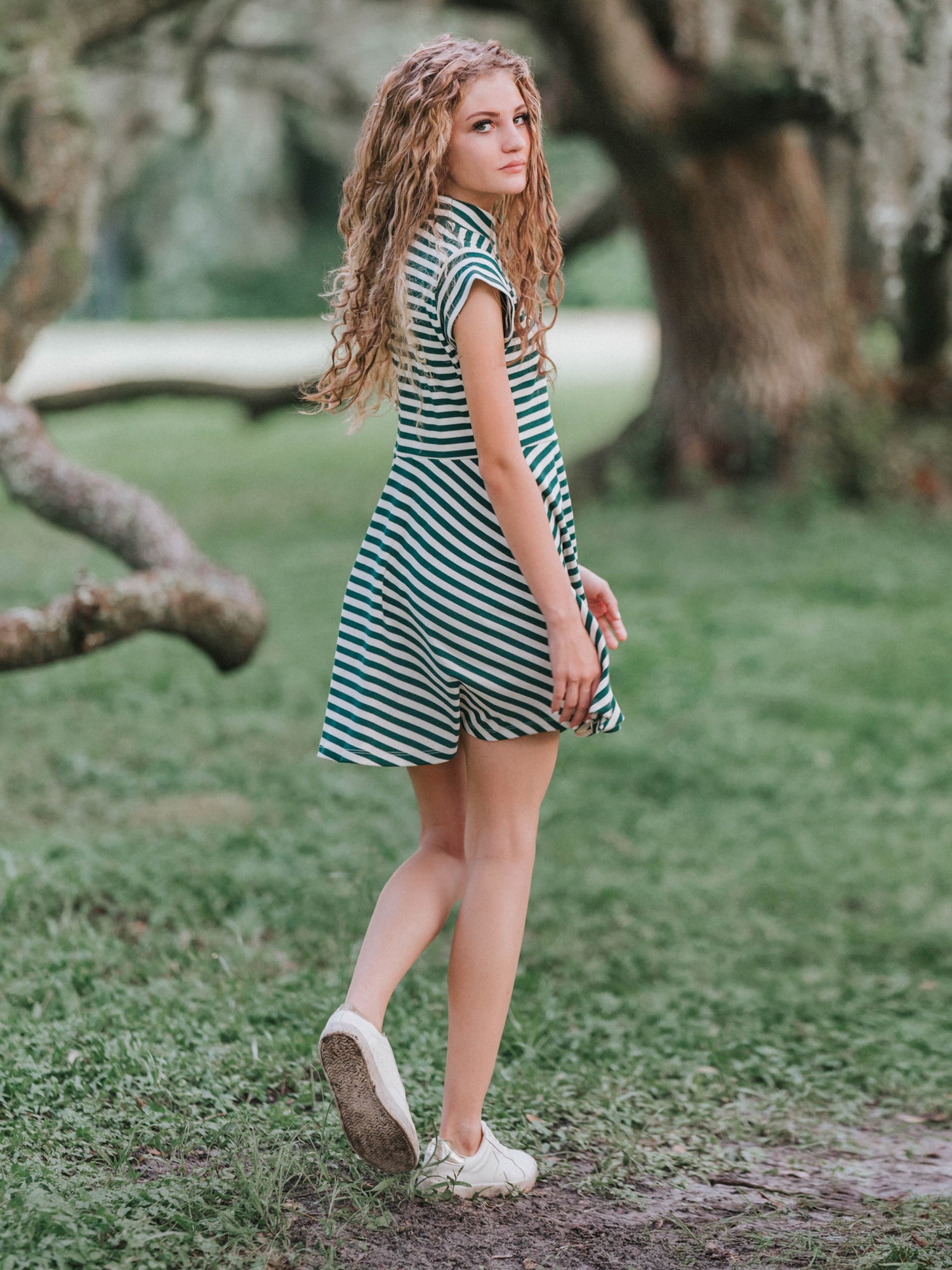 Girl outside in a greenery area smiling looking at the camera wearing a deep green and ivory striped dress. Model is also wearing white canvas shoes. 