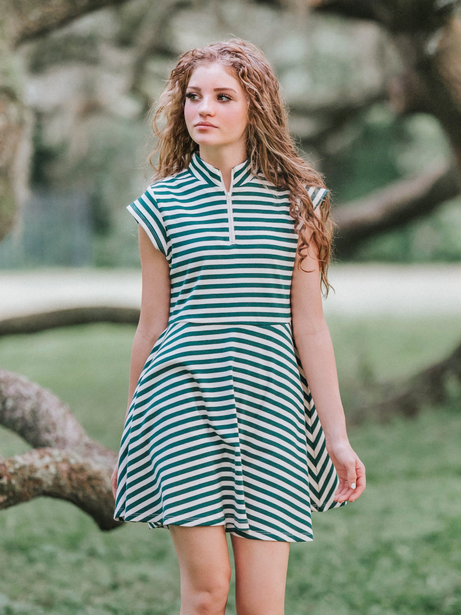 Girl outside in a greenery area smiling looking at the camera wearing a deep green and ivory striped dress.
