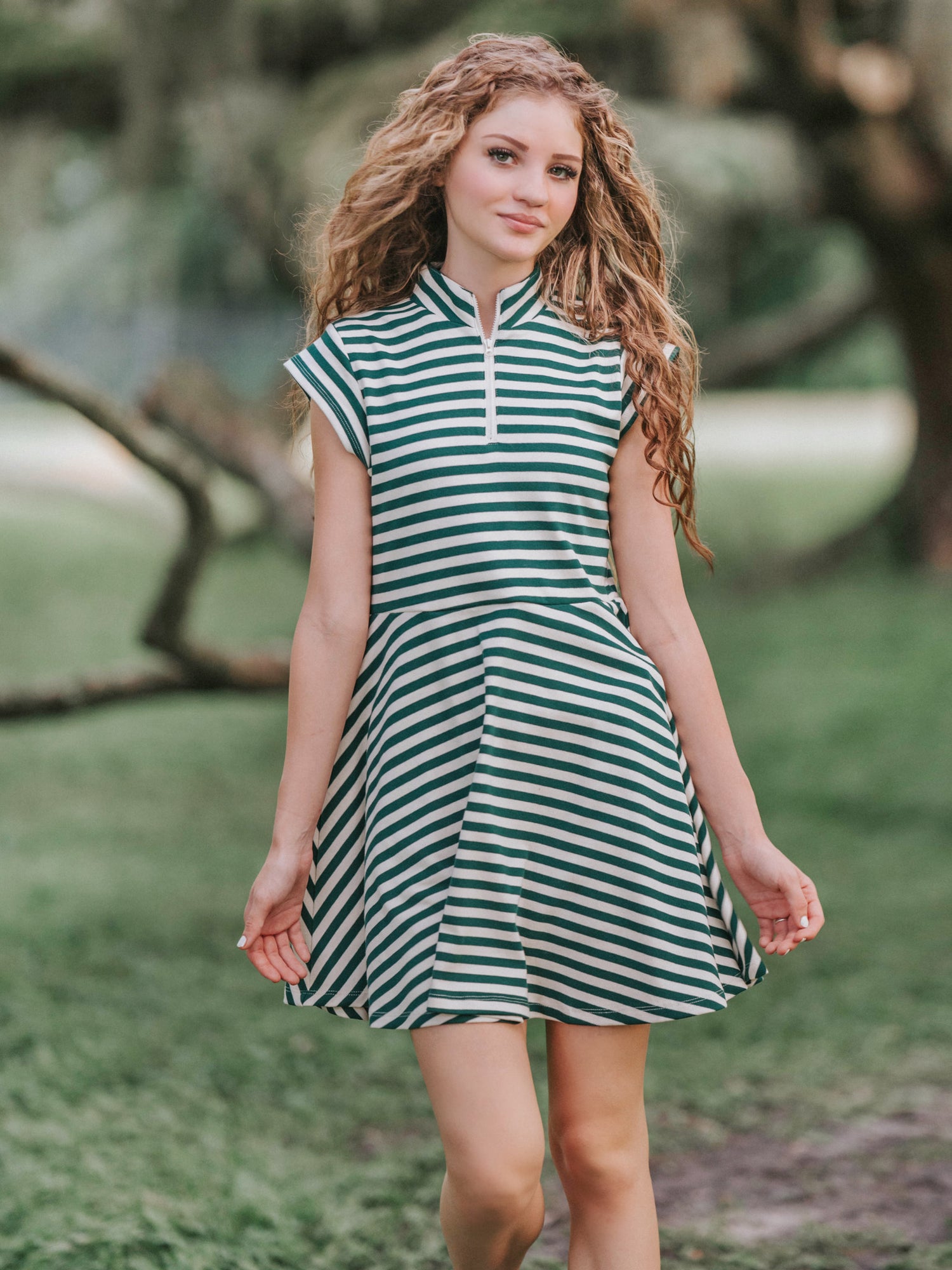 Girl outside in a greenery area smiling looking at the camera wearing a deep green and ivory striped dress.