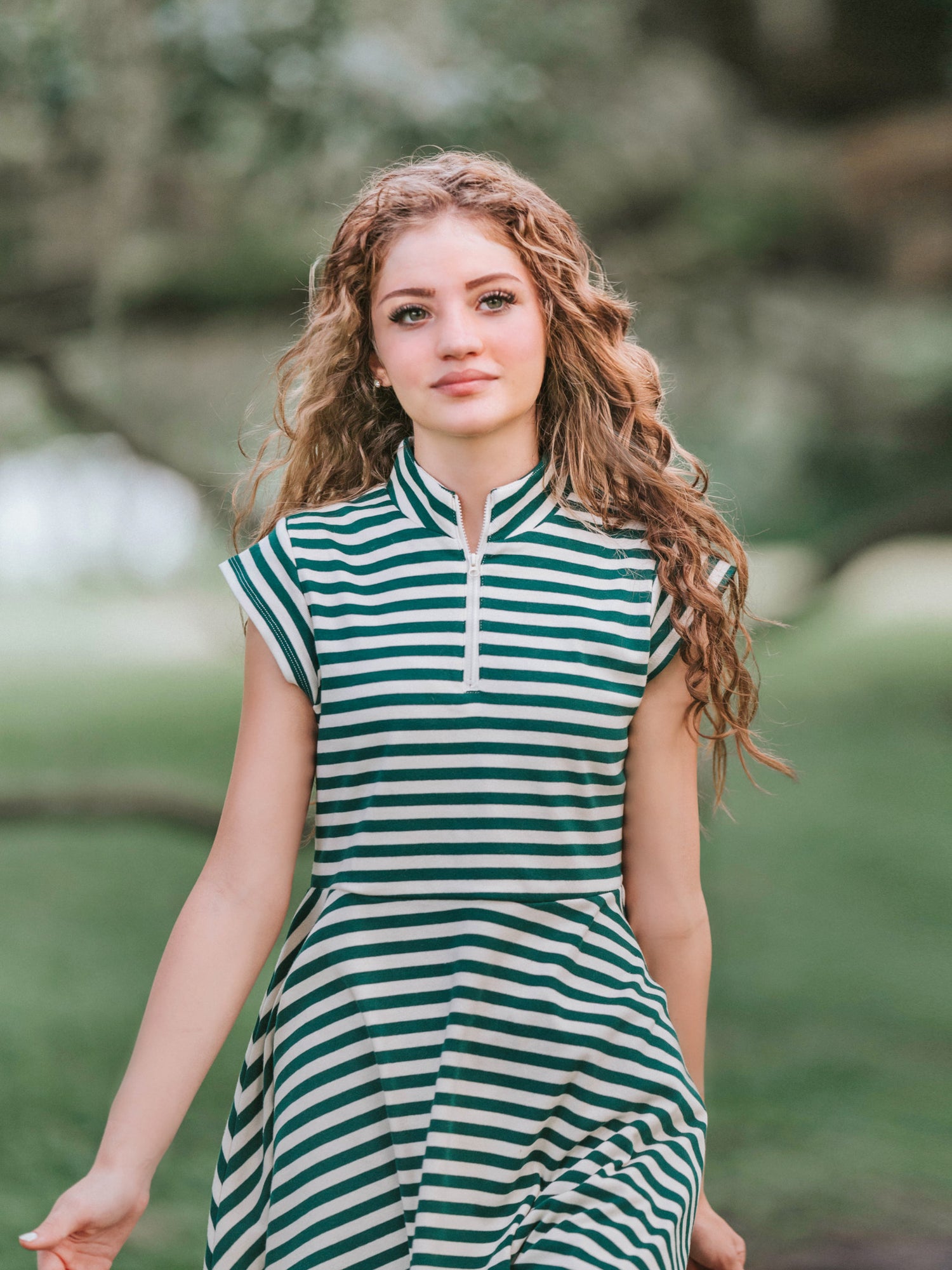 Girl outside in a greenery area smiling looking at the camera wearing a deep green and ivory striped dress.