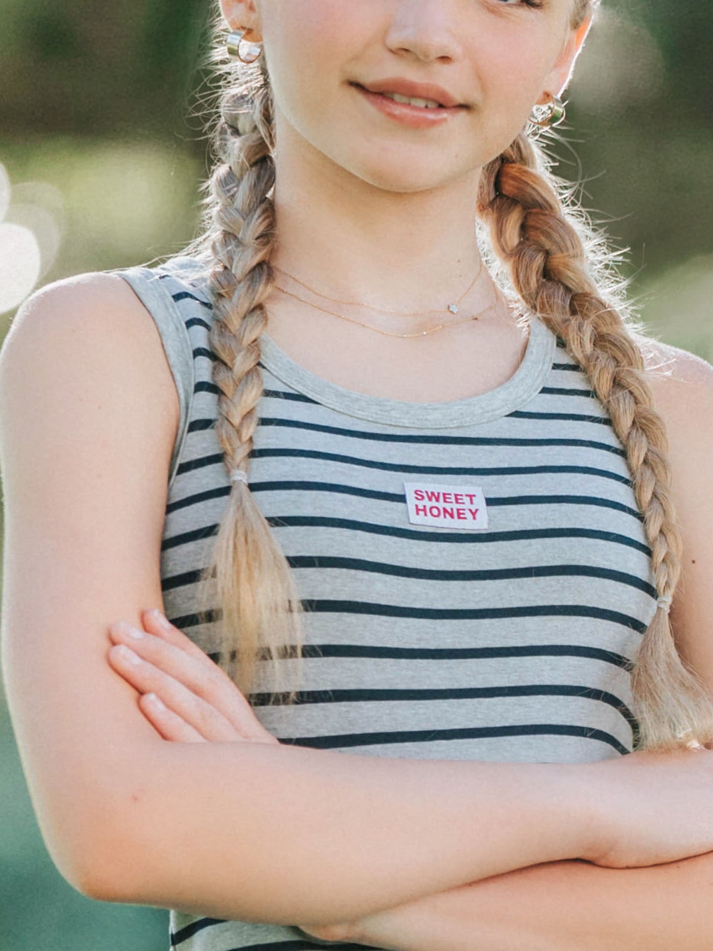 Up close picture of a model wearing a grey and navy striped sleeveless shirt. There's a sweethoney logo tag in the center of the chest and the model has arms folded. 
