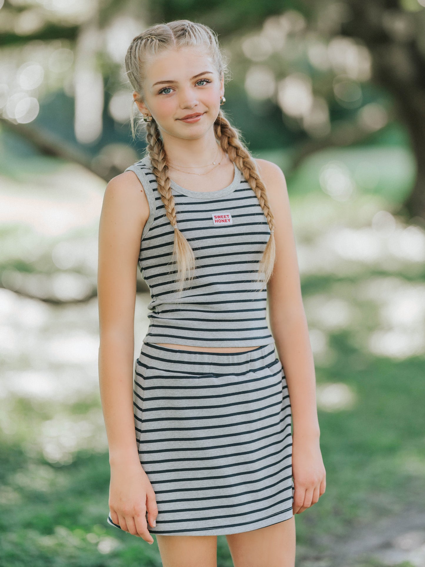 Model is outside and is wearing a two piece skort set that is grey and navy striped. Shirt is sleeveless and has a sweethoney logo tag in the middle of the chest. 