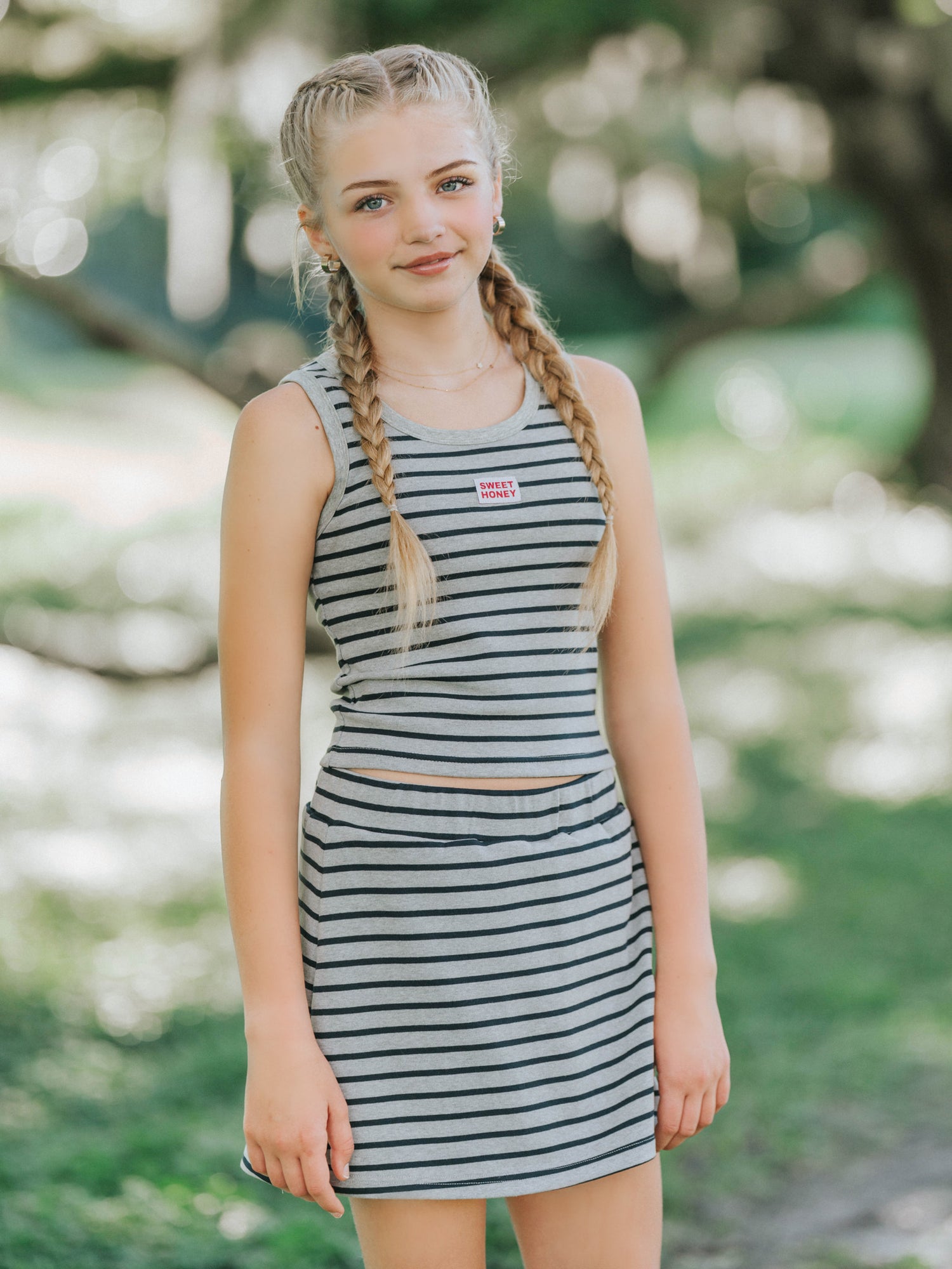 Model is outside and is wearing a two piece skort set that is grey and navy striped. Shirt is sleeveless and has a sweethoney logo tag in the middle of the chest. 
