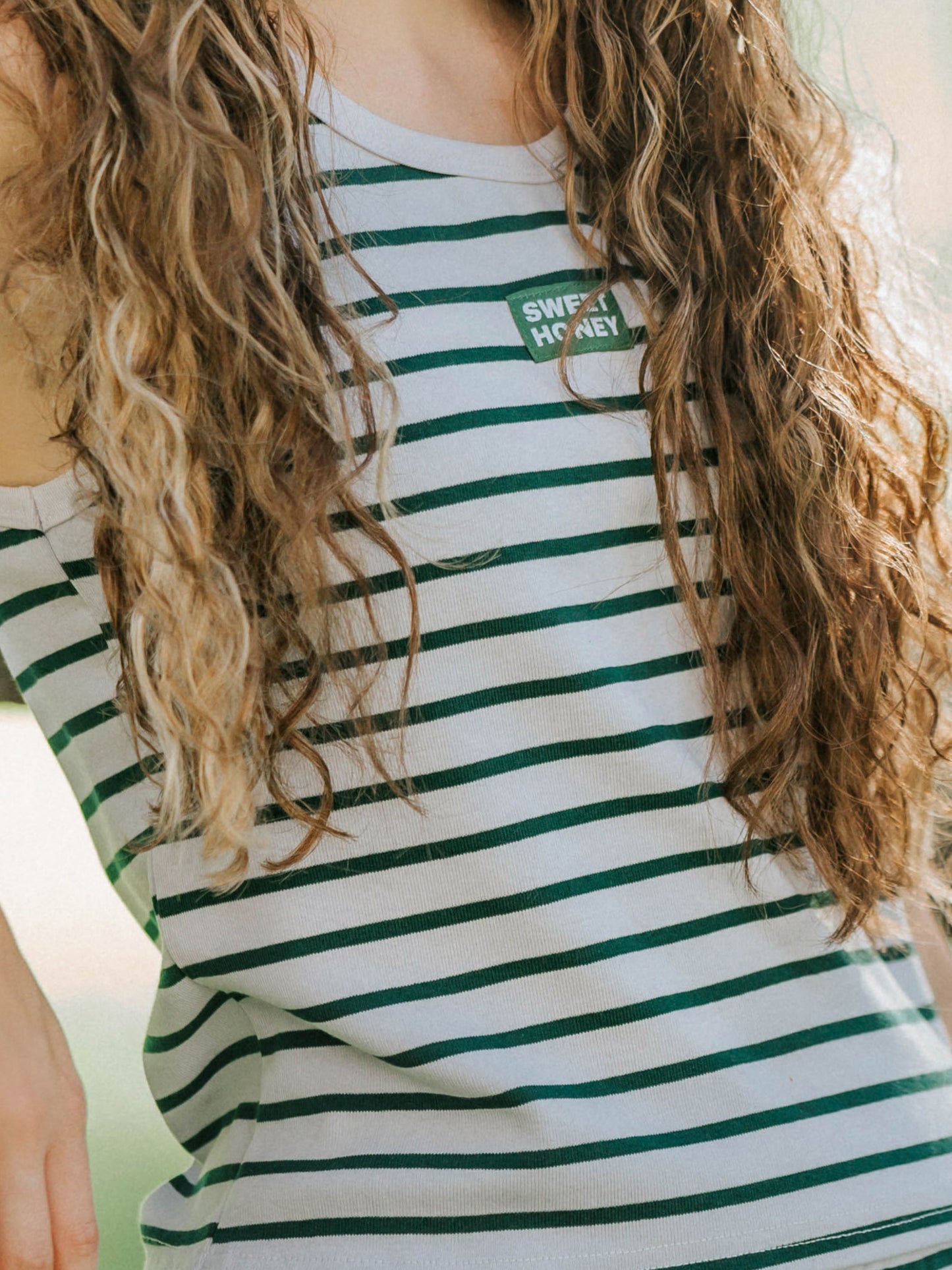 Up close picture of a model wearing a grey and green striped tank top. In the middle of the chest of the shirt is a SweetHoney logo tag. 