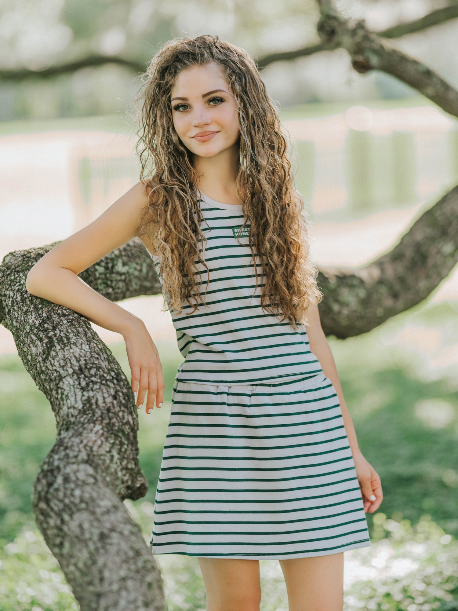 Model is outside and has her elbow propped on a tree branch. Model is wearing a two-piece skort set that is greyish and green striped. Shirt is sleeveless. 