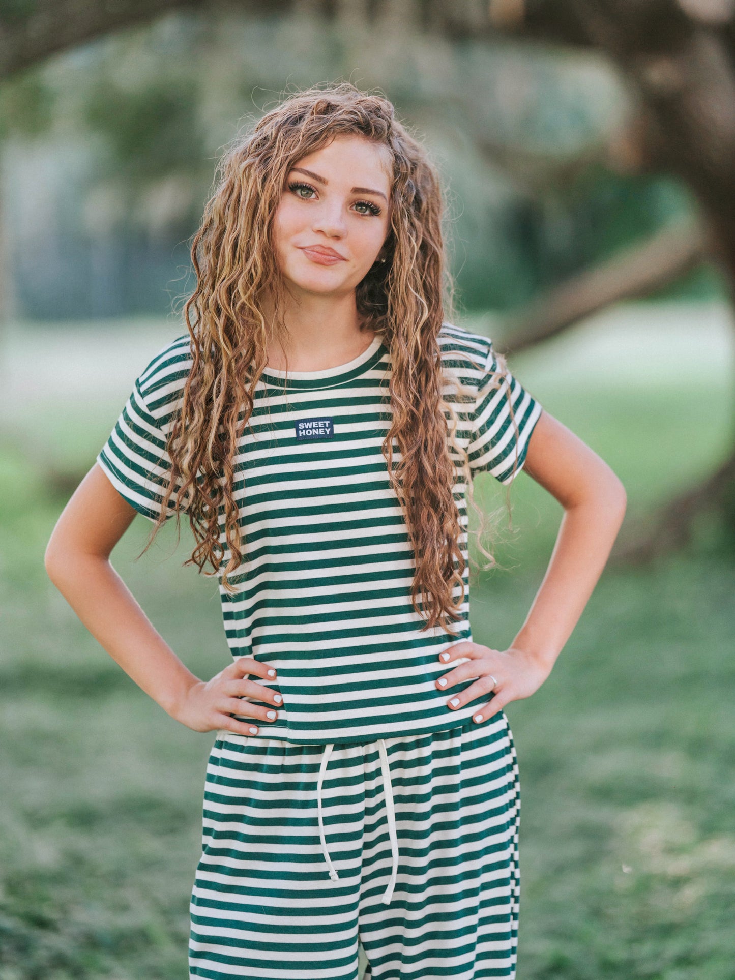 Model is in a mossy, greenery area and is wearing a two piece hunter green and ivory striped set. Model has hands on her hips. 