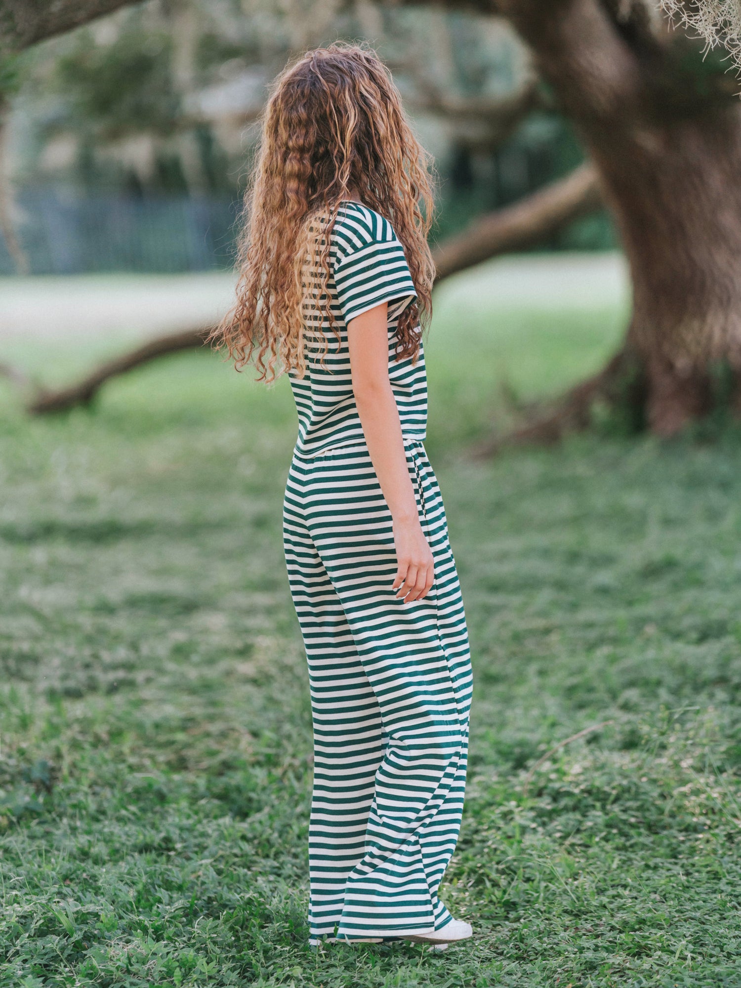 Model is outside in a mossy area and she is wearing a two piece hunter green and ivory pant set. 