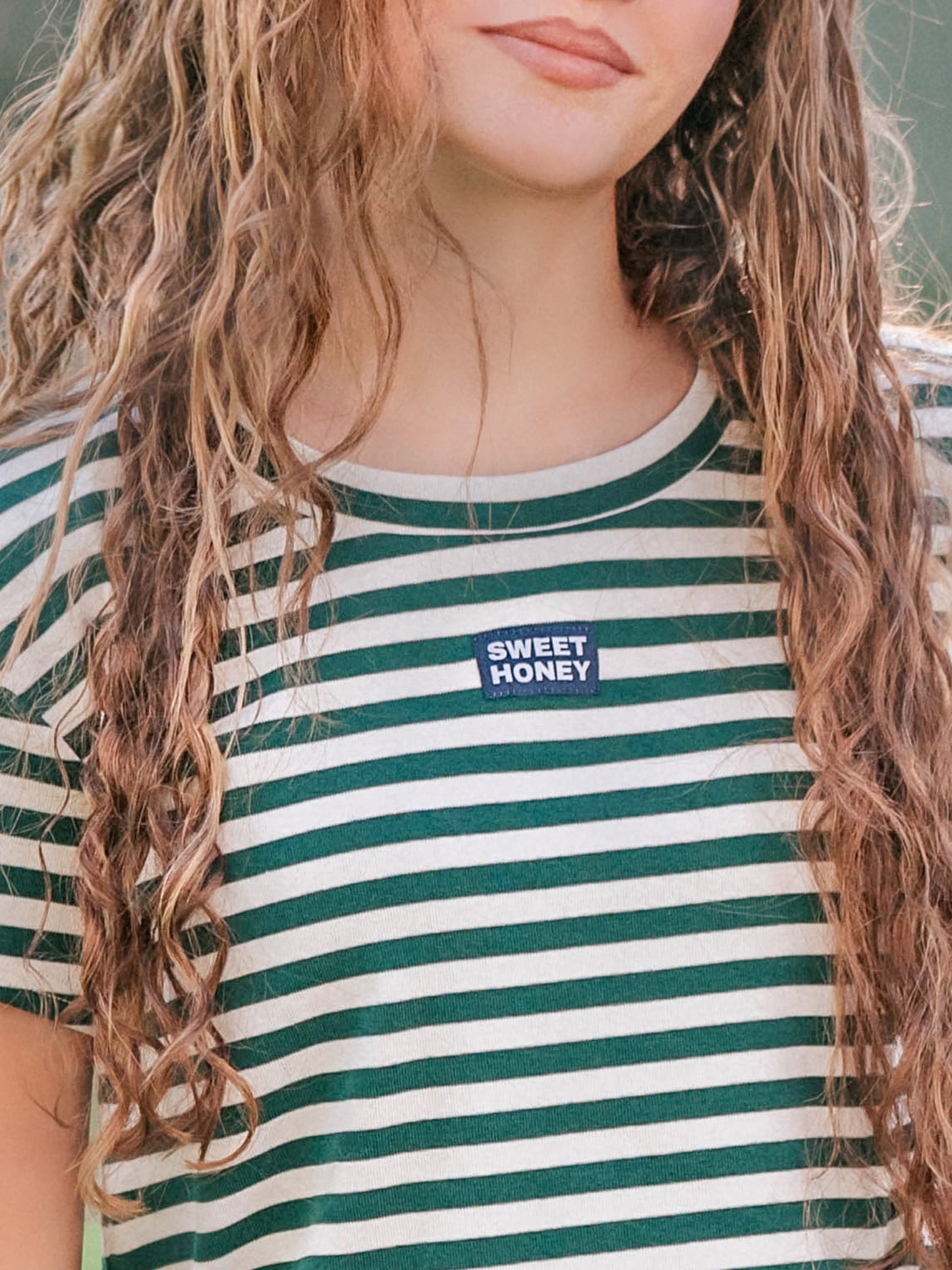 Close up of a green and ivory striped shirt. Shirt has a SweetHoney logo tag in the middle of the chest. 
