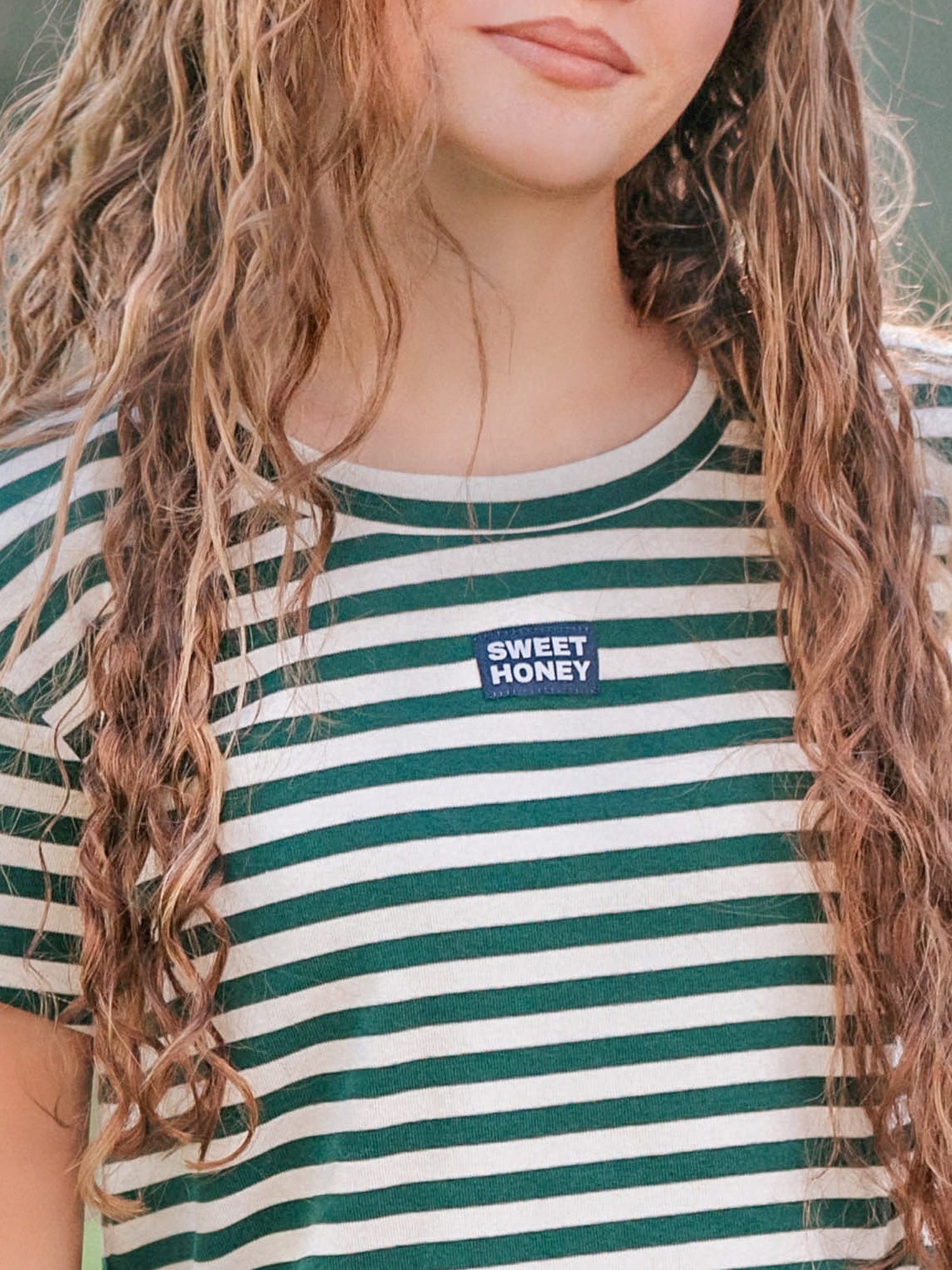 Close up of a green and ivory striped shirt. Shirt has a SweetHoney logo tag in the middle of the chest. 