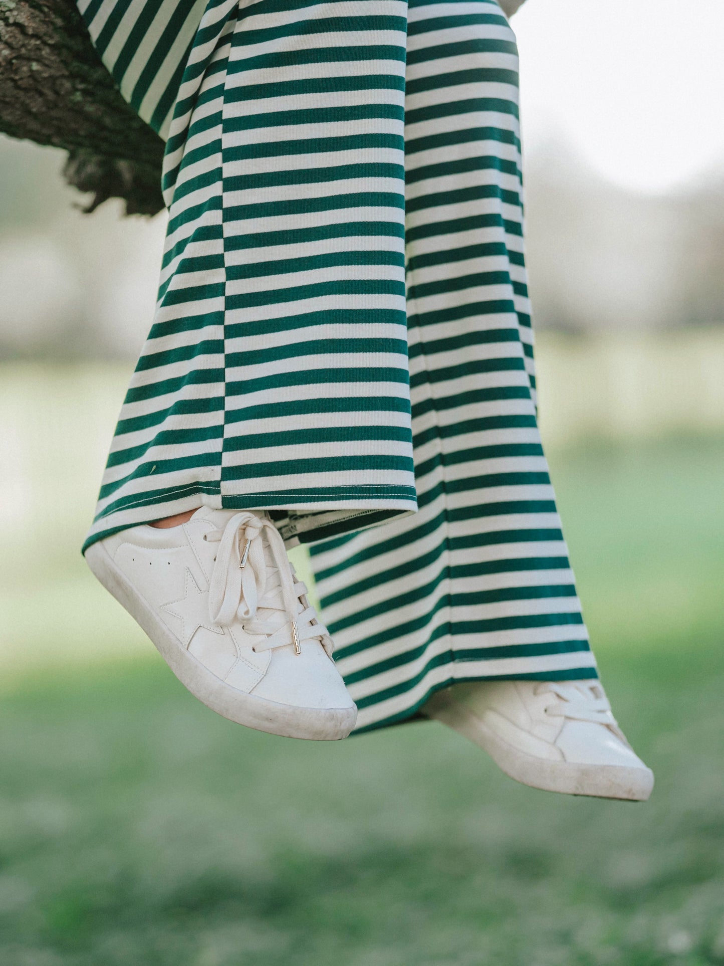 Model is in a mossy, greenery area and is wearing a two piece hunter green and ivory striped set. Models legs are dangling and she is wearing white canvas shoes. 