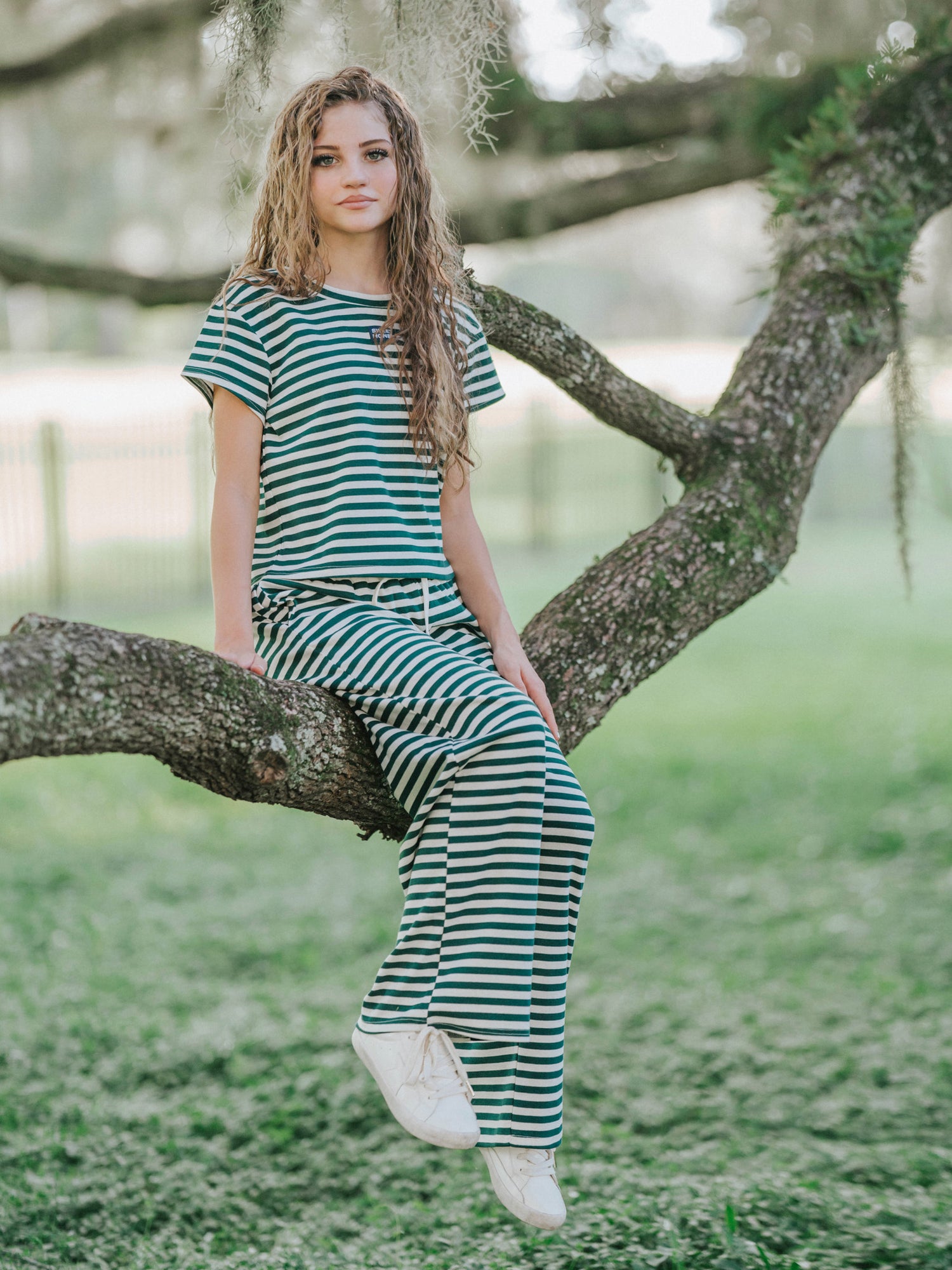 Model is in a mossy area sitting on a tree branch and is wearing a hunter green and ivory two piece striped set. 