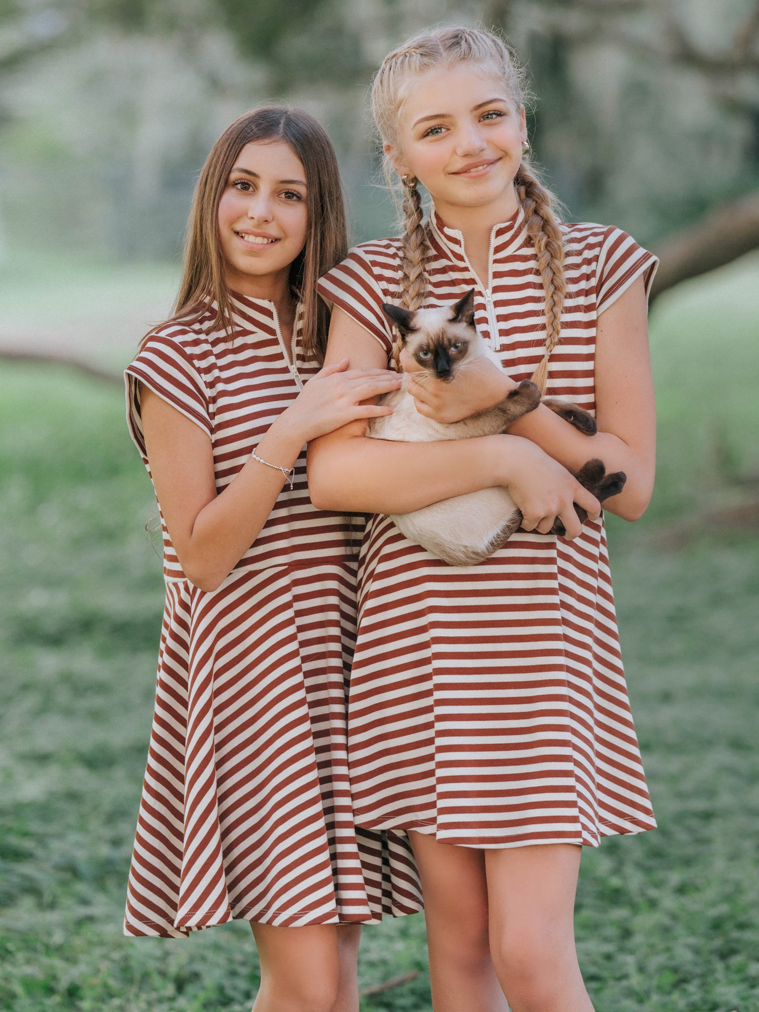 Two models outside and one model is holding a persian cat. Both models are wearing matching dresses that are brown and ivory striped with cap sleeves. 