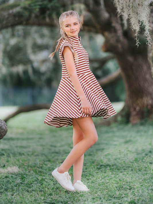 Model is outside in a green area wearing a chocolate and ivory striped dress that has cap sleeves and a zipper at chest. Model is slowly twirling and wearing white shoes. 