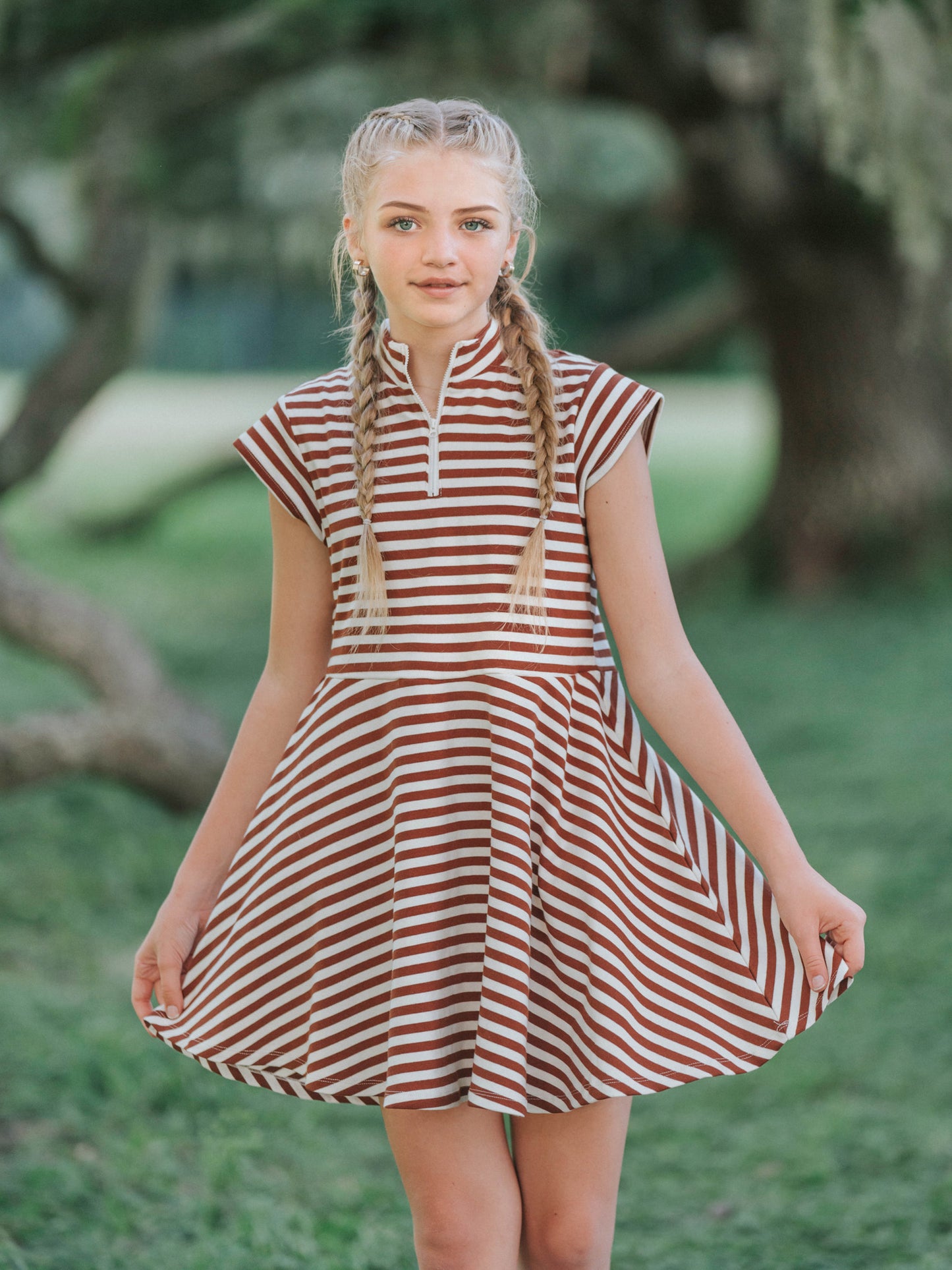 Model is outside in a green area wearing a chocolate and ivory striped dress that has cap sleeves and a zipper at chest. Model is holding the sides of dress out. 