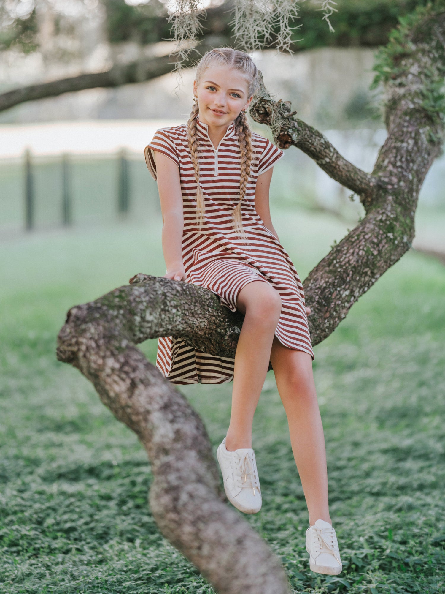Model is outside sitting on a tree branch. Her feet are dangling and she is wearing a chocolate brown dress with cap sleeves and a zipper on chest. Model has white canvas shoes on.