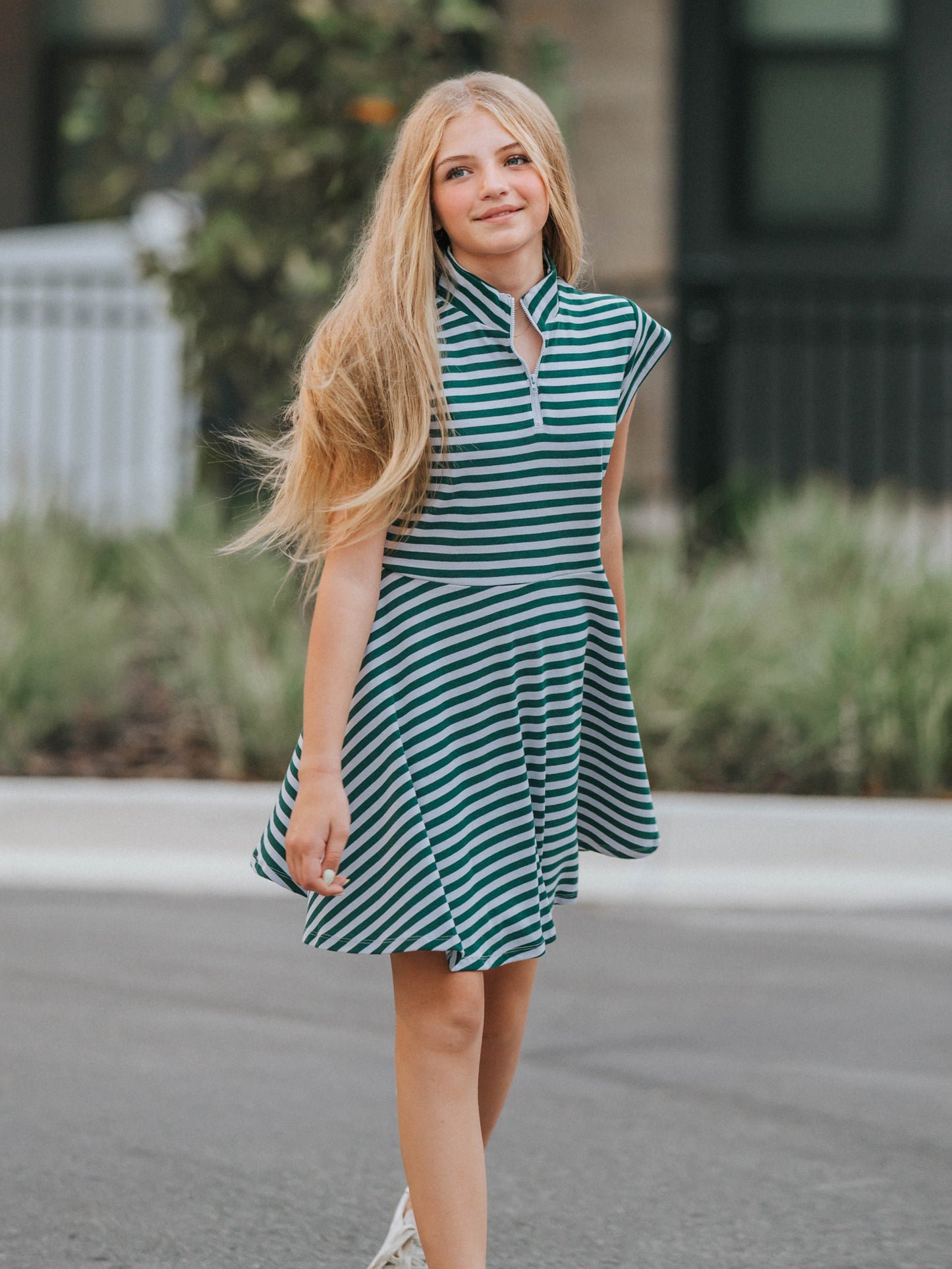 Model is outside in street wearing a lavender and green stiped dress with white canvas shoes. 