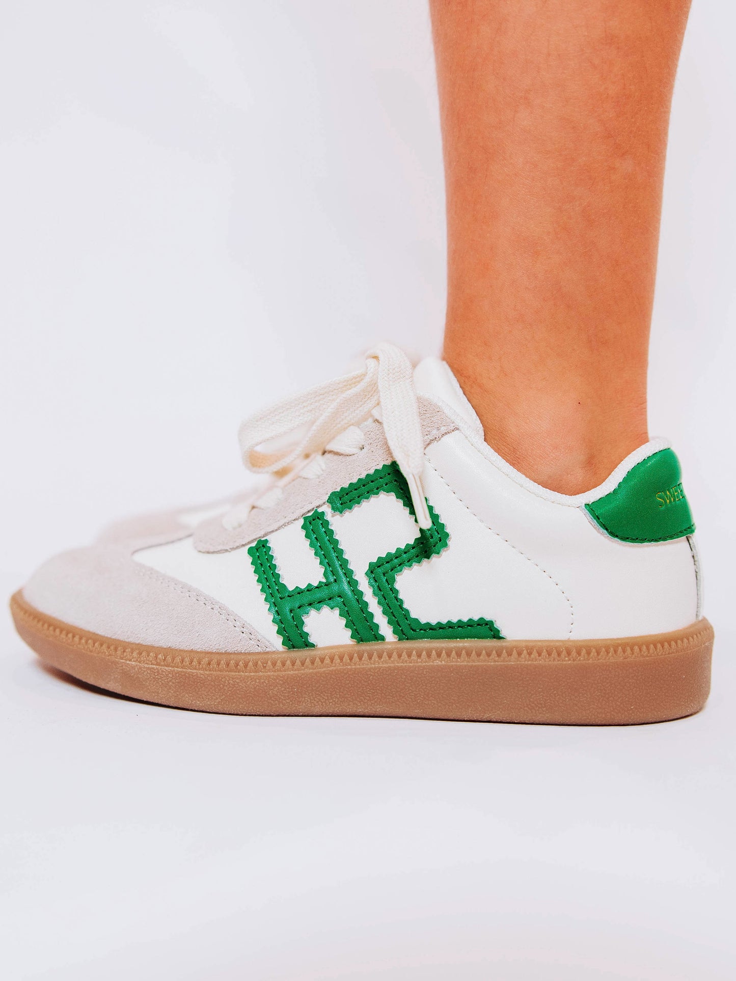 Left shoe side view of girl wearing white sneakers with "SH" lettering stitched in green leather on outer side of shoes along with green heel caps.