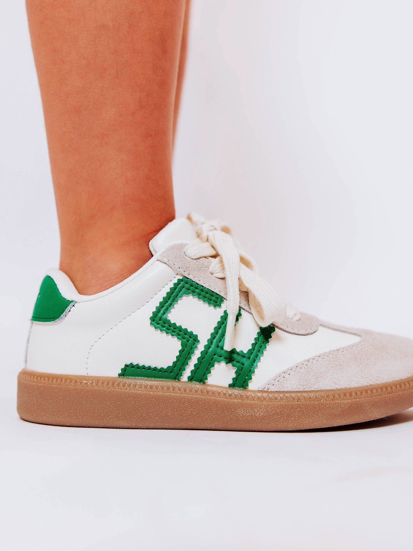 Close shot of side view of girl wearing white sneakers with "SH" lettering stitched in green leather on outer sides of shoes along with green heel caps.