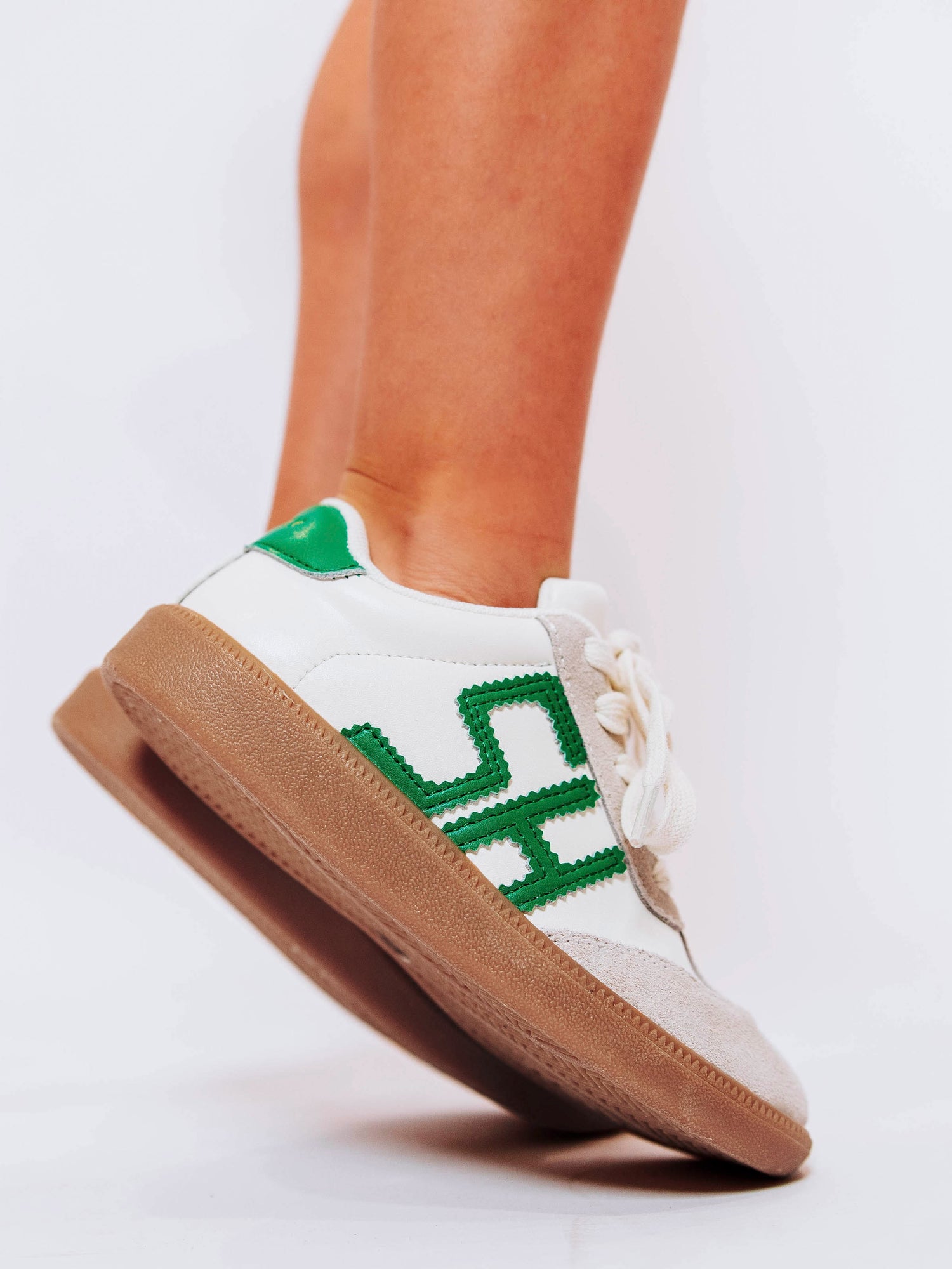 Close shot of girl standing on her toes wearing white sneakers with "SH" lettering stitched in green leather on outer sides of shoes along with green heel caps.