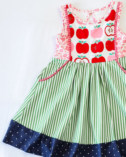 Flay lay of  a dress with main apple pattern on bodice and green striped skirt with white polka dotted blue hem