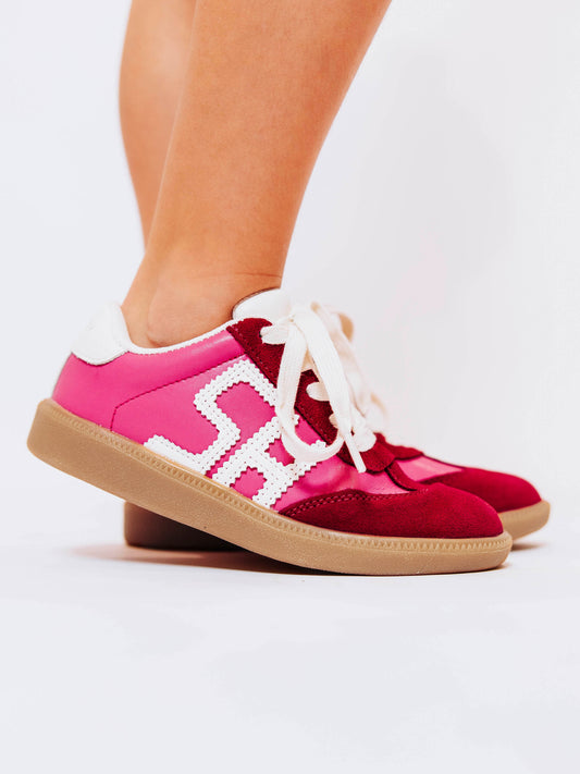 Close shot of girl wearing pink sneakers with red suede secondary details and "SH" lettering in stitched white leather on the outer side of the shoes.