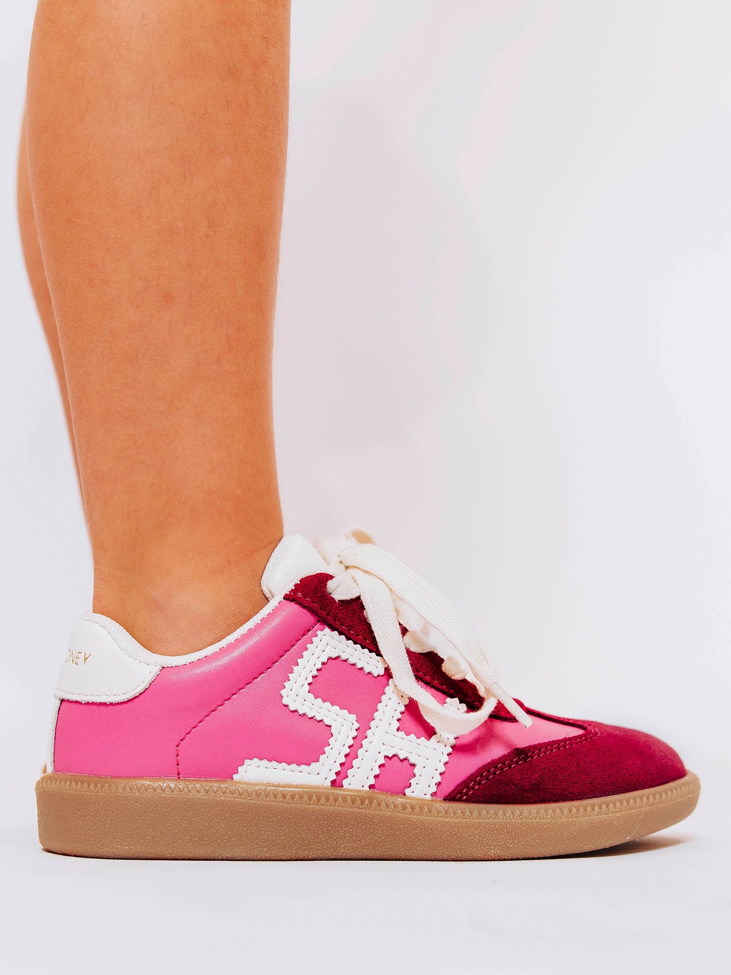 Close shot of side view of girl wearing pink sneakers with red suede secondary details and "SH" stitched in white leather on the outer side of the shoes.