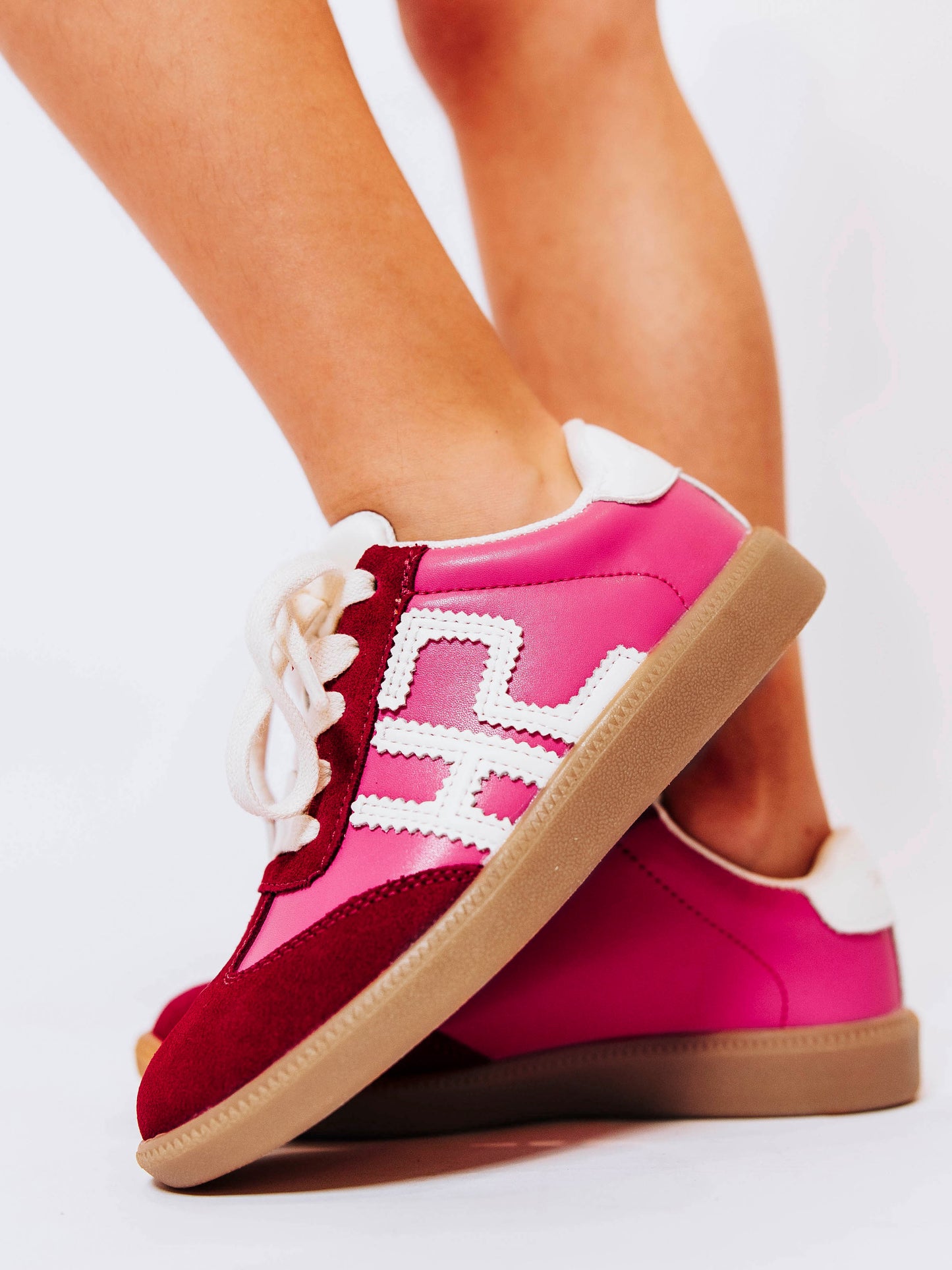 Close shot of side view of girl wearing pink sneakers with red suede secondary details and "SH" in stitched white leather on the outer side of the shoes.