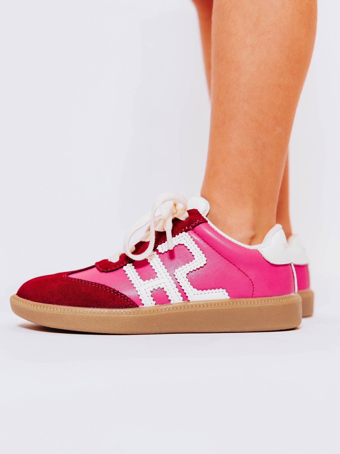 Side view of girl wearing pink sneakers with red suede secondary details and "SH" in white stitched leather on the outer side of the shoes.