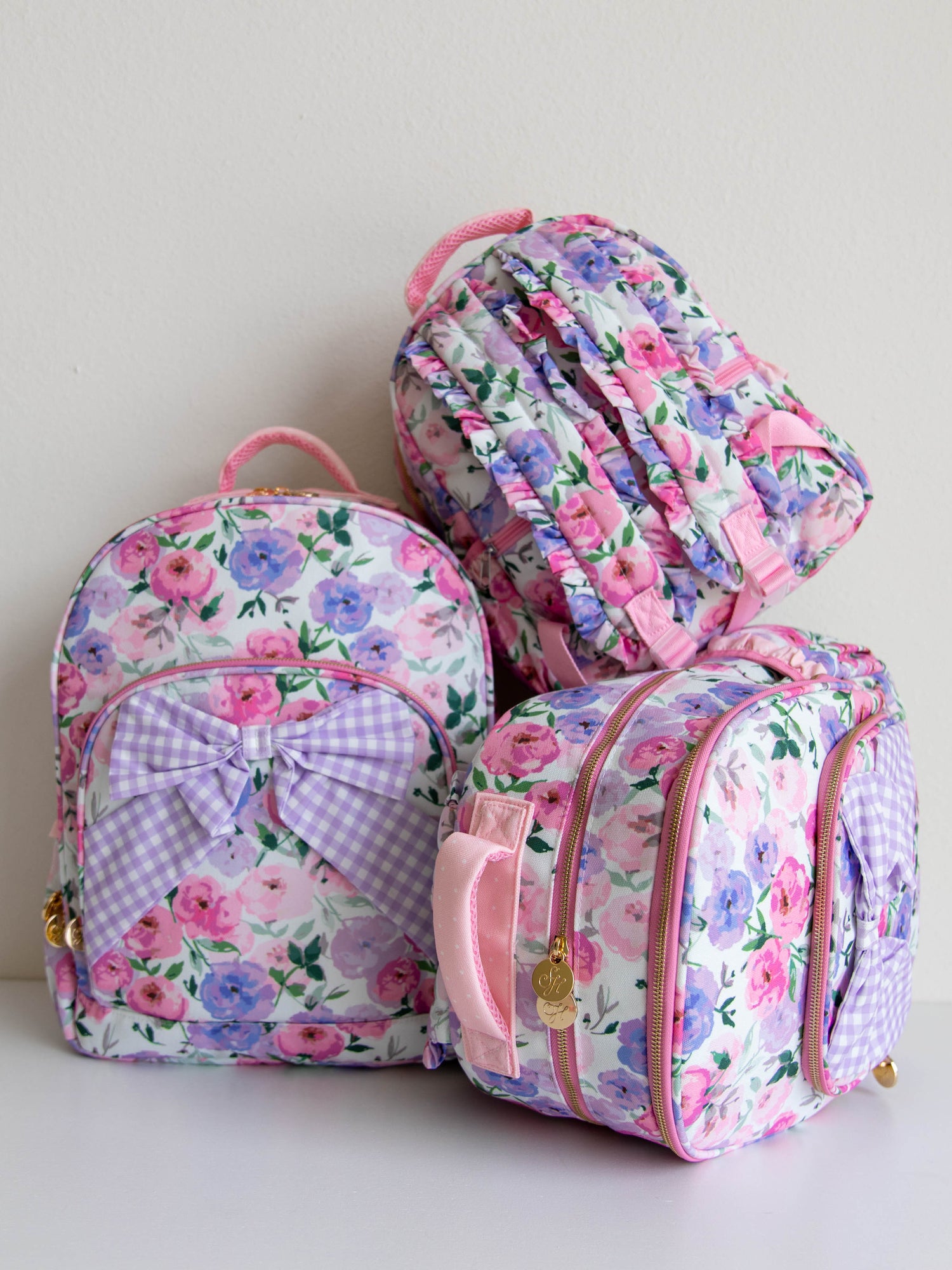 Different size stacked  colorful floral backpack with a purple gingham bow attached to the backpack. gold zippers and zipper pull
