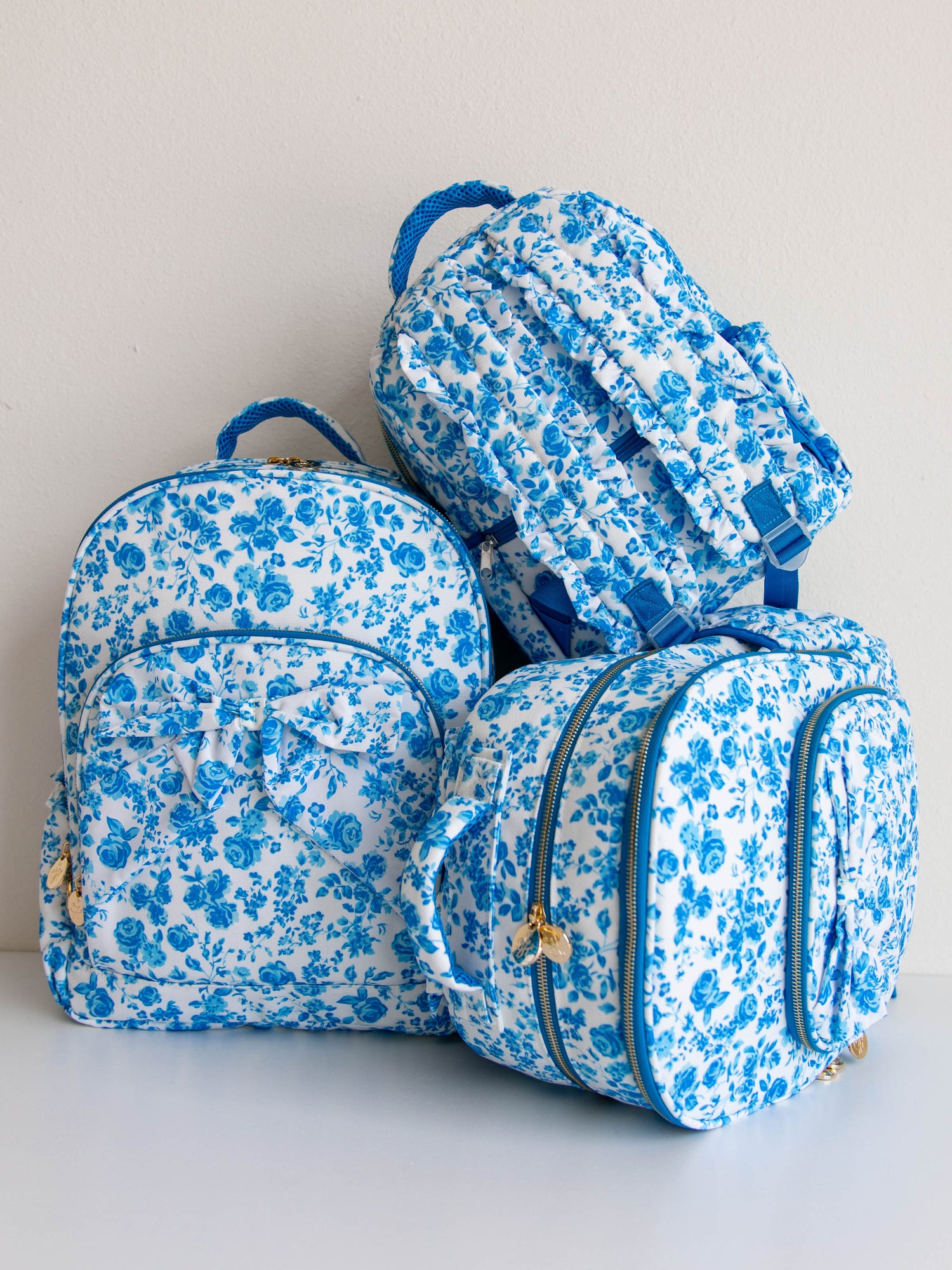 Picture of three different sized blue, floral backpacks. All with gold zipper pulls and attached bows on the front
