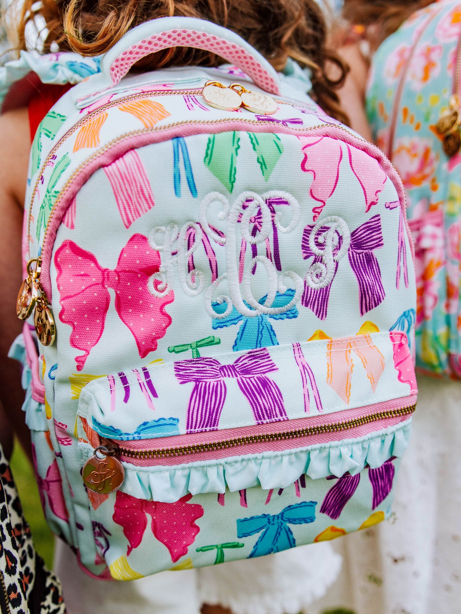 Girl wearing a Vibrant bows printed backpack with customized embroidery, gold zippers and zipper pulls, and a handle