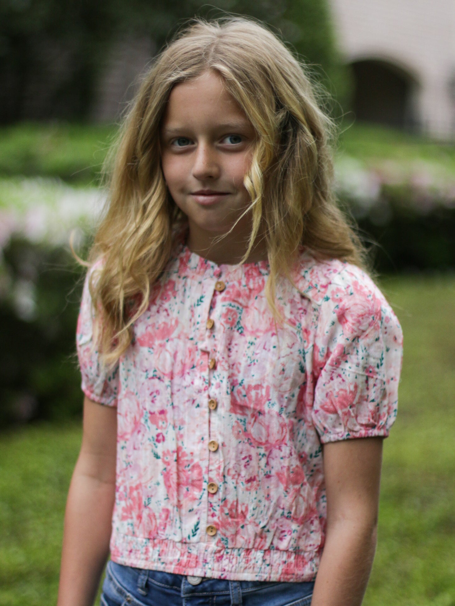 This image of a girl features the product Classic Button Top - Starburst Pink. This top has functional buttons down the bodice, puff sleeves, and elastic ruffle neckline. It is a vibrant pattern of large bursting pink blooms and small green leaves.