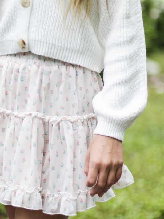 This image of a girl features the product Classic Tiered Mini Skirt – Floral Drop. This double layered short skirt features a smocked waistband as well as ruffled seams and hem. It is a pattern of tiny hand drawn flowers spaced out across a background of cream.
