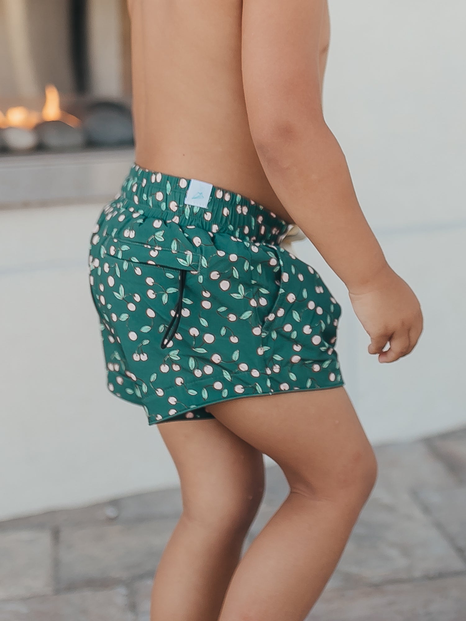 This image of a boy features the European cut style of our Boy's Everyday Lined Trunks – Cherry on Top. This pair of trunks is a pattern of light pink cherries on a dark green background.