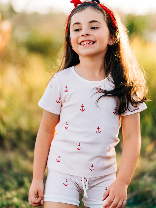 This image of a girl features the product Everyday Shorts Set – Anchors in Red. This ribbed knit set of short sleeve shirt and shorts comes in a pattern of red anchors on an almost white, pink background.