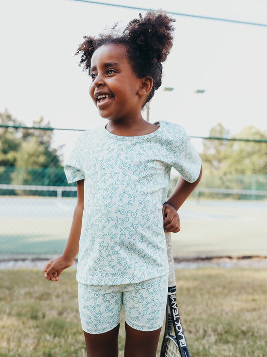 This image of a girl playing tennis features the product All in Motion Set – Teal Petals. This activewear set of a shirt and shorts is a pattern of teal line drawn flowers and leaves over an almost white background.