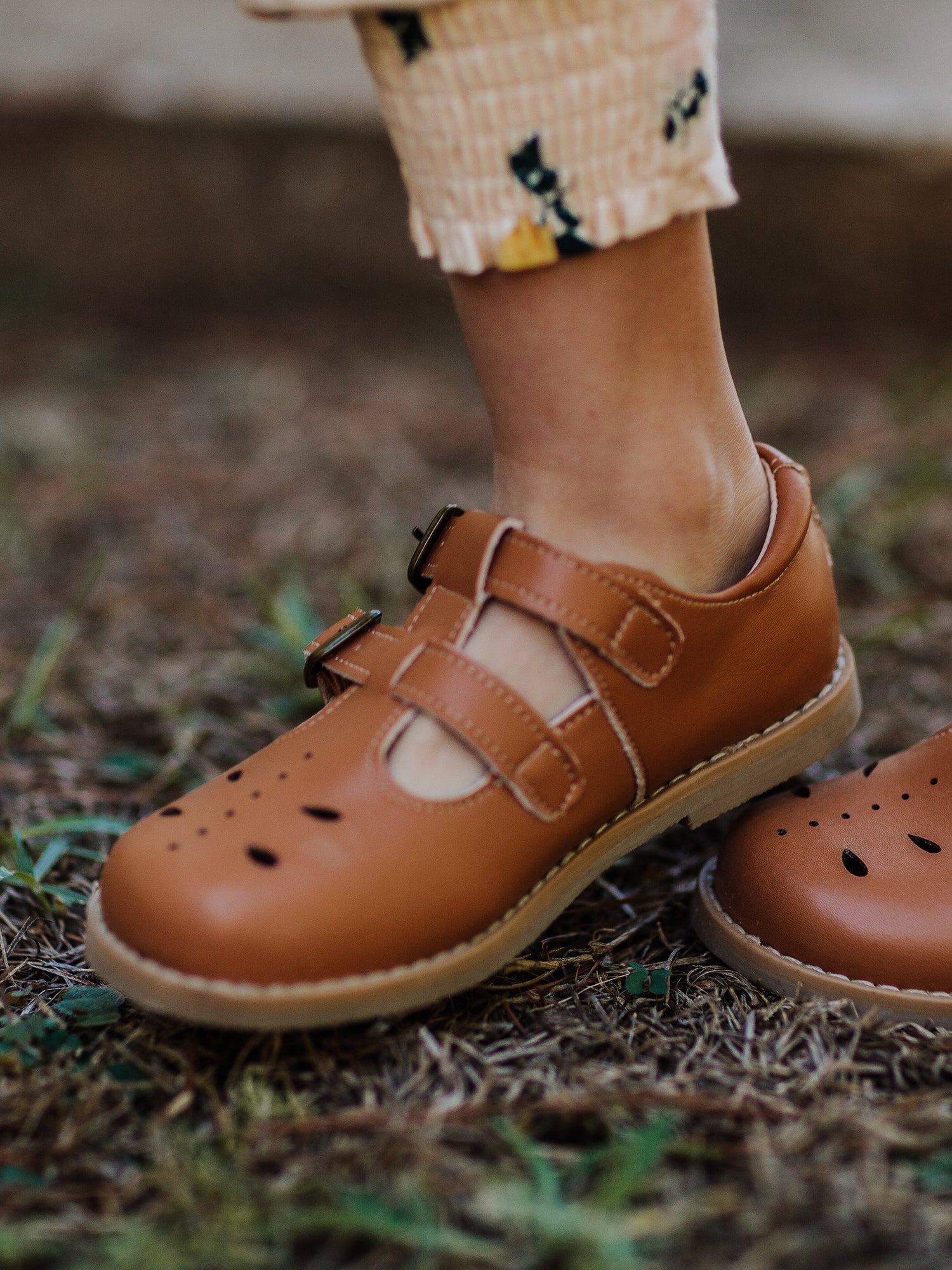 A pair of camel colored genuine leather rubber soled shoes with two adjustable metal buckles and ventilation holes across the toe area in a dragonfly type pattern.