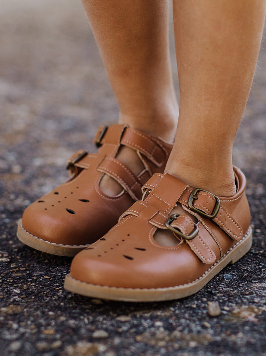 A pair of camel colored genuine leather rubber soled shoes with two adjustable metal buckles and ventilation holes across the toe area in a dragonfly type pattern.