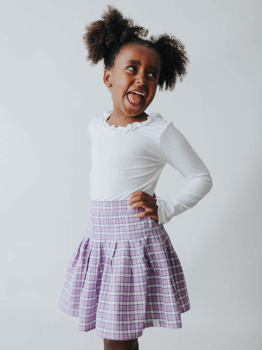 This image of a girl features the product Pleated Skort – Purple Check. This pleated skirt comes with built-in shorts and an elastic back in a check pattern of purple, navy, and white.