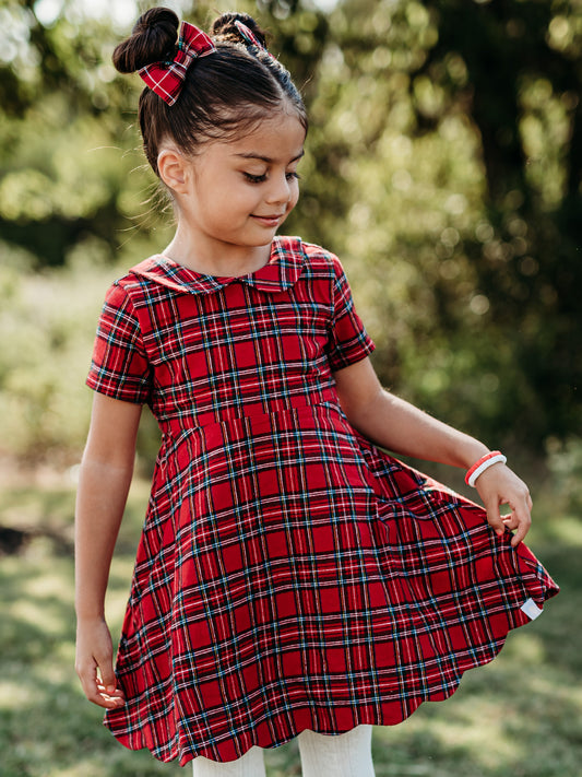 This image of a girl features the product Retro Peter Pan Dress - Holiday Red Plaid. It comes in a red plaid pattern with accents of white, green, blue, and yellow.