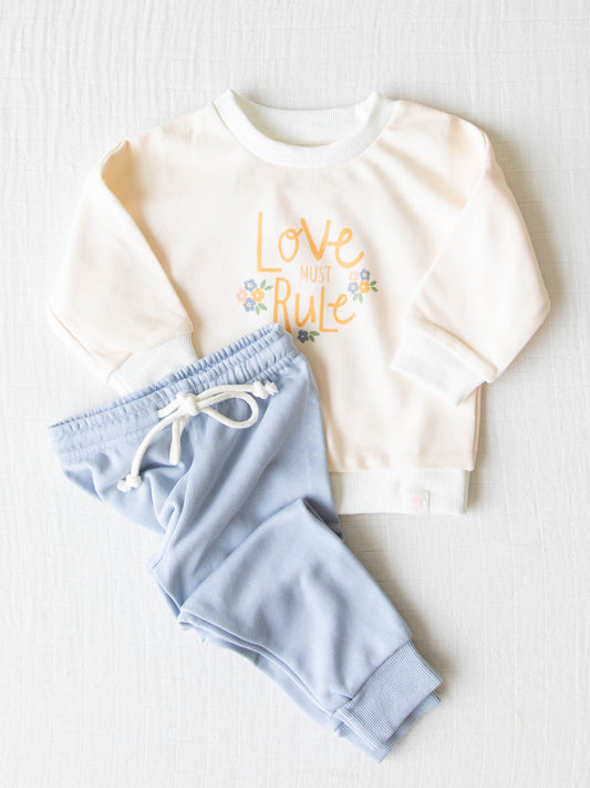 Sweatshirt Set - Love Must Rule. This set has ribbed cuffs and an elastic waistband with functional drawstring. The top has “Love Must Rule” on it with little clusters of flowers on a cream background. The bottoms are a light blue color.